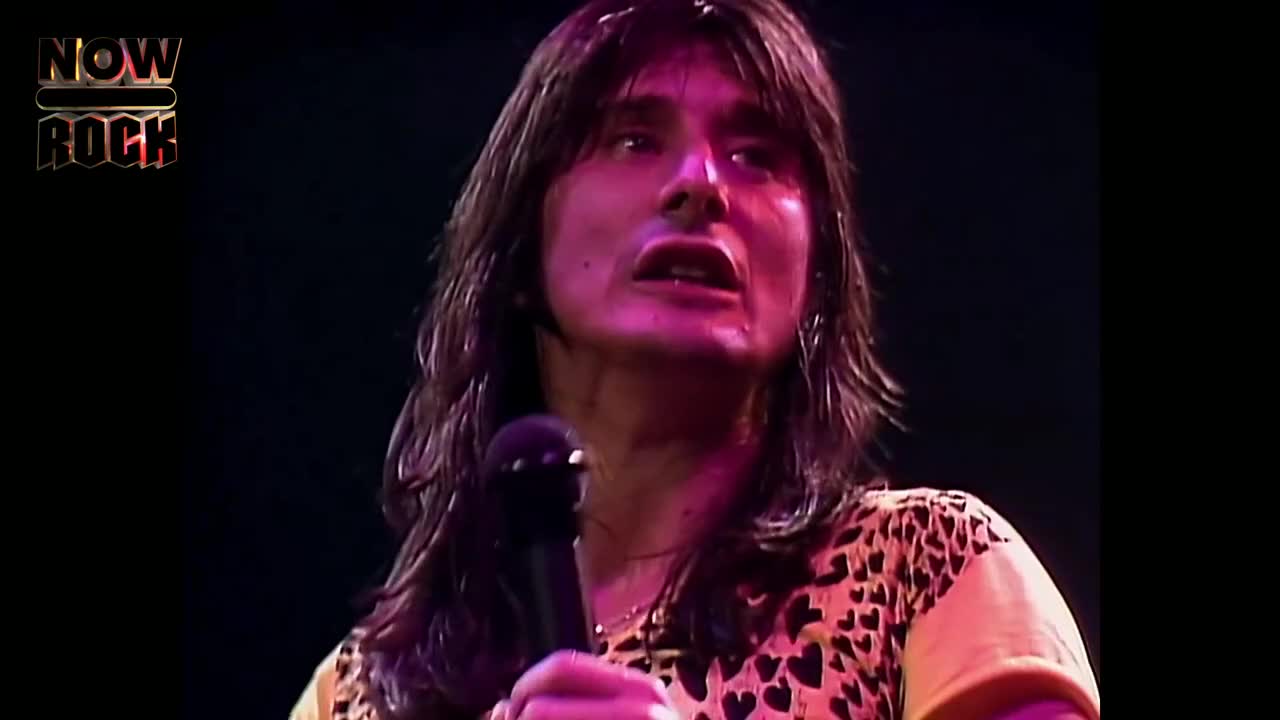 The singer, mic in hand, is mid-song, eyes wide and mouth open. His long hair hangs over his shoulders as he belts out a tune, illuminated by the stage lights.
