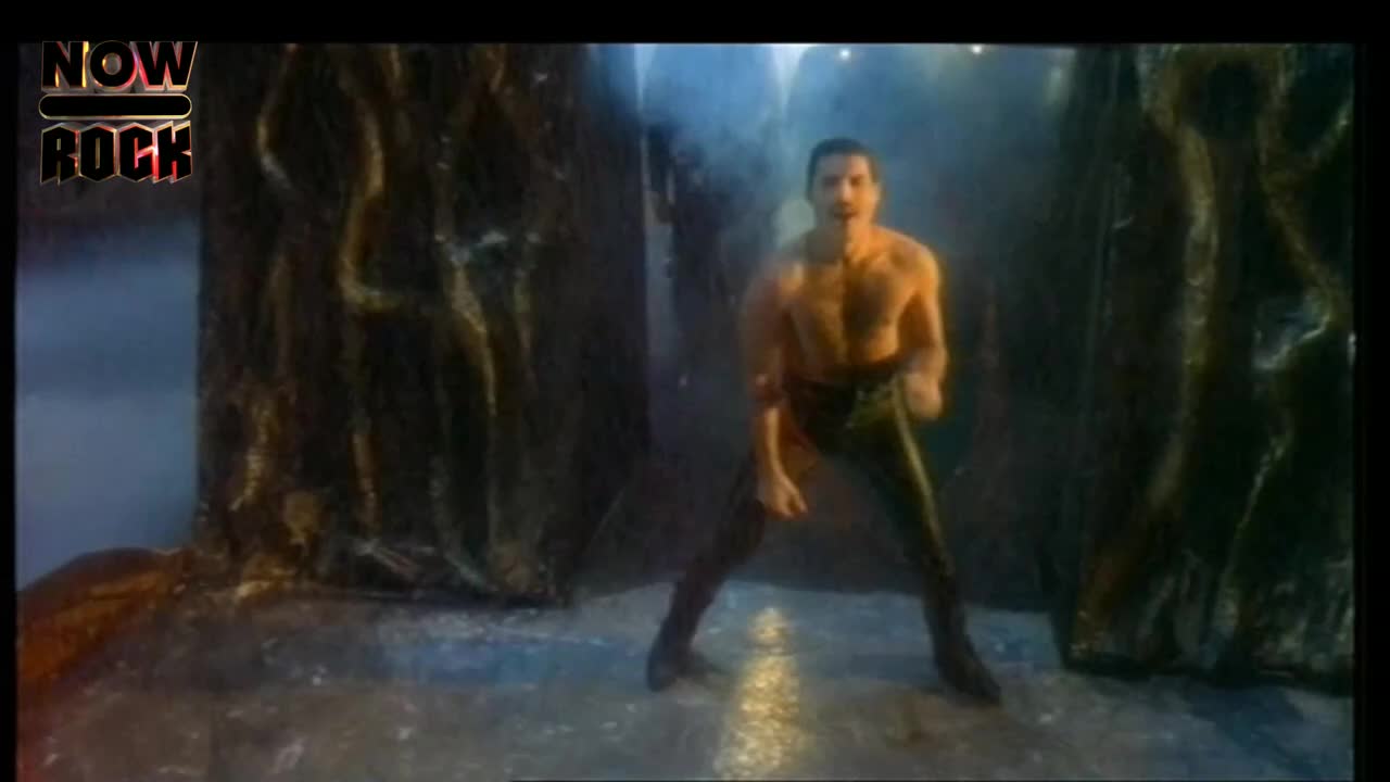 He's center stage, bare-chested and in tight black pants, striking a pose. The set looks like a cave, and the lighting gives everything a dramatic, almost ethereal, feel.
