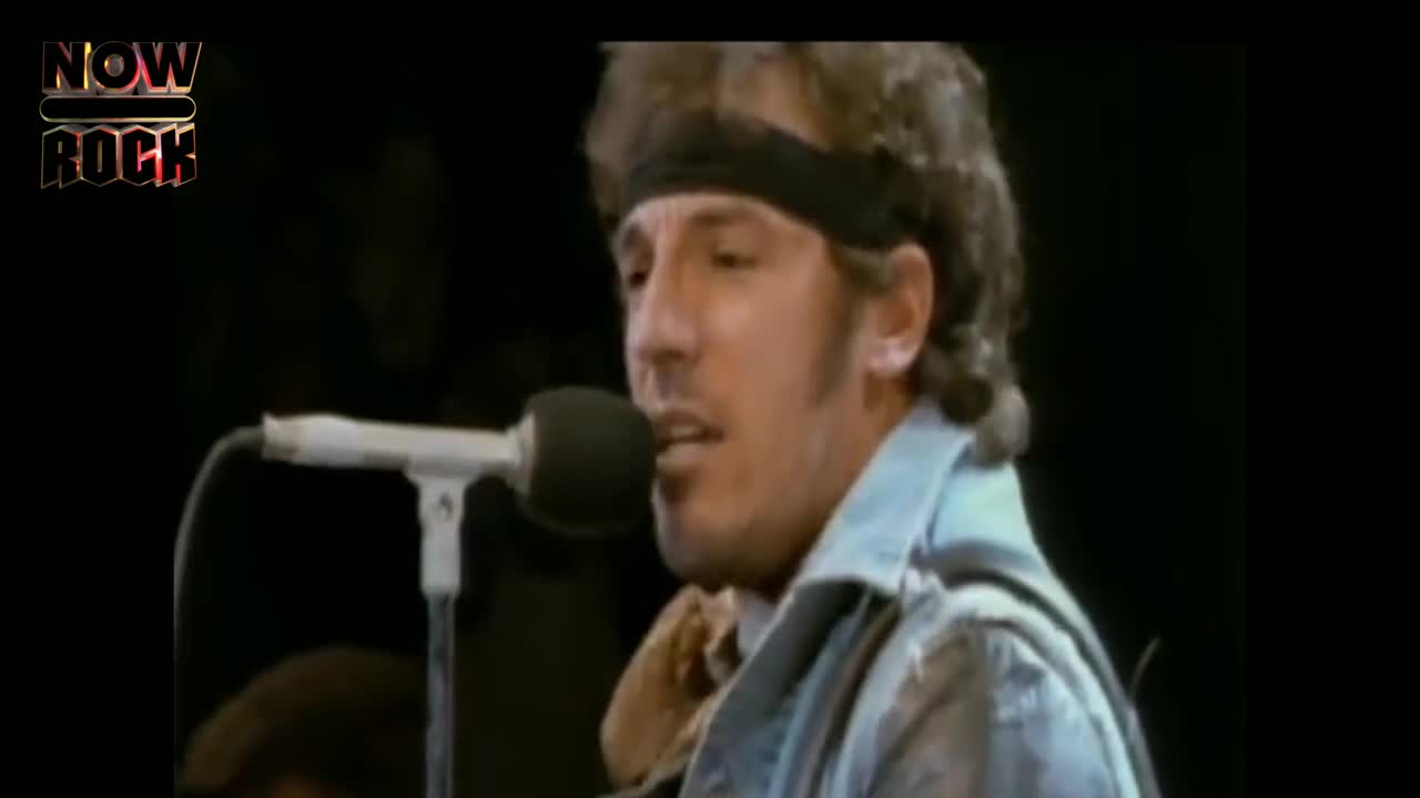 He's singing into a microphone, his face illuminated by the stage lights. A black headband keeps his hair out of his eyes as he belts out the lyrics.
