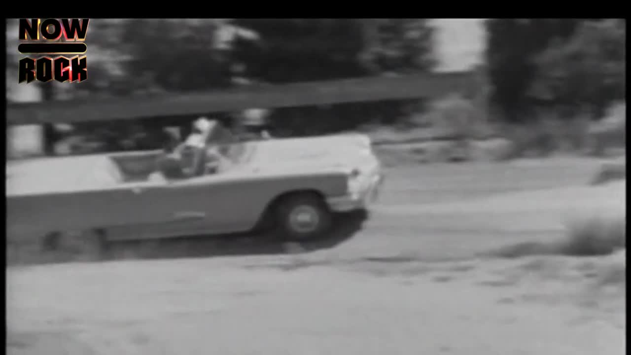 A convertible, its top down, kicks up dust as it speeds along a dirt road. The car is moving fast, and the scene is a bit blurry.
