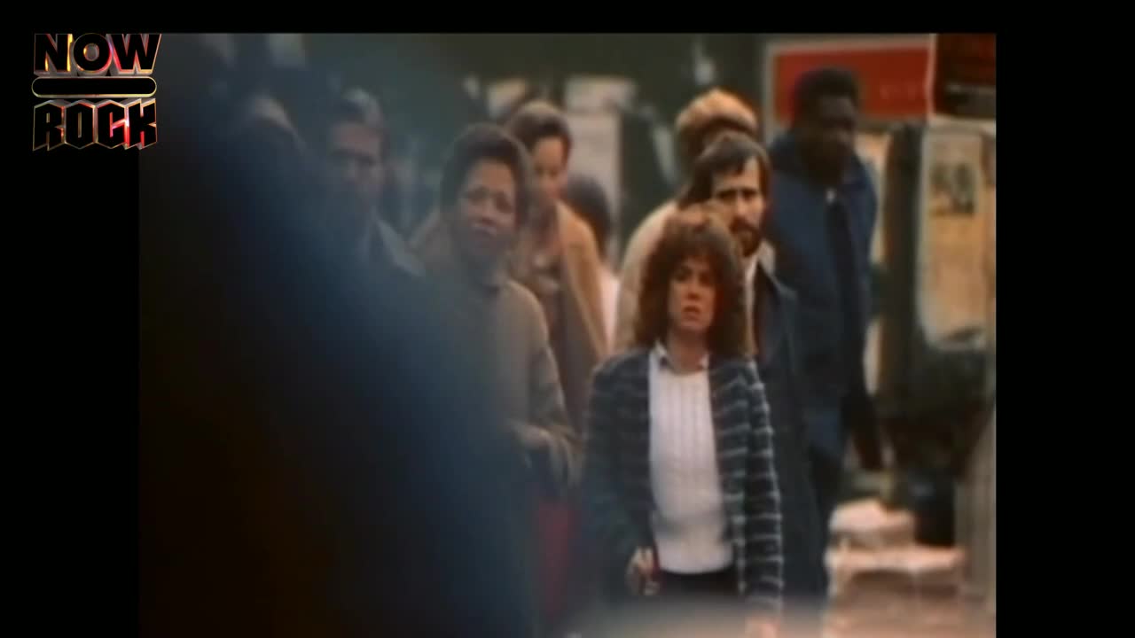 People are walking on a busy street, the woman in the striped jacket and white shirt is looking directly ahead. The "NOW ROCK" logo is in the upper left corner.
