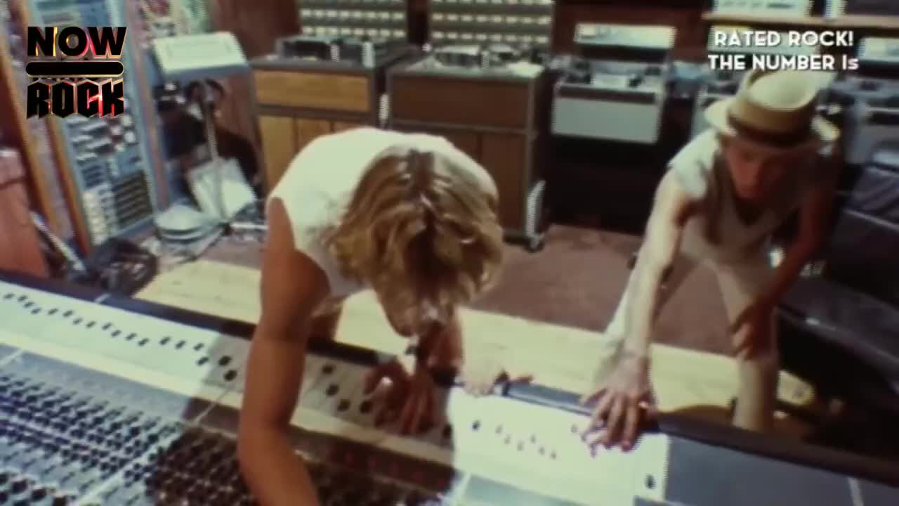 Two guys lean over a mixing console, their hands hovering over the controls. The one on the right wears a hat, and they both seem focused on something.
