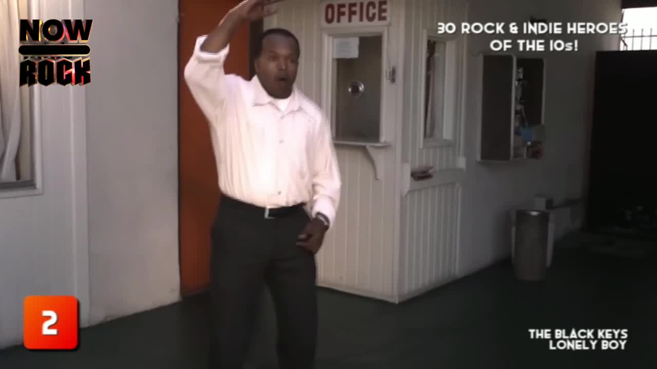 A man in a white shirt raises his arm, gesturing dramatically near a small office window. The NOW Rock logo and a list of "Rock & Indie Heroes" are visible in the background.
