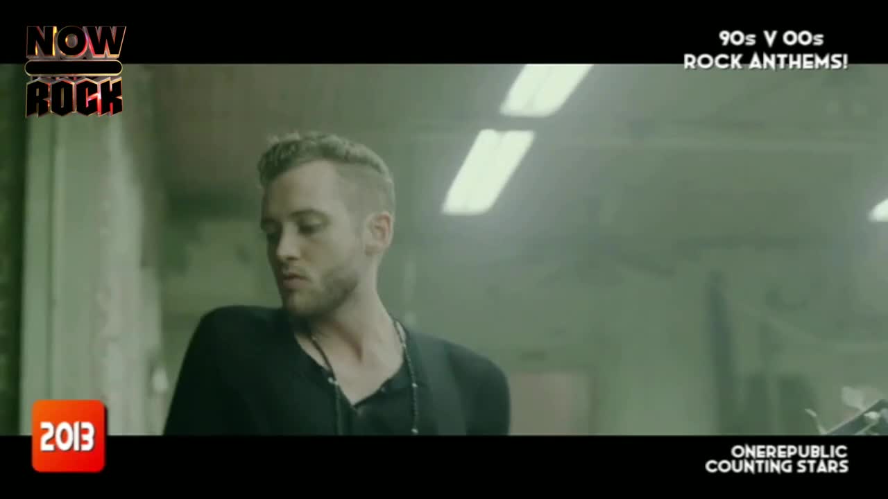 A man with a close-cropped haircut is looking down, holding a guitar out of frame. The scene is lit with a muted, hazy light, like an old music video from the UK.
