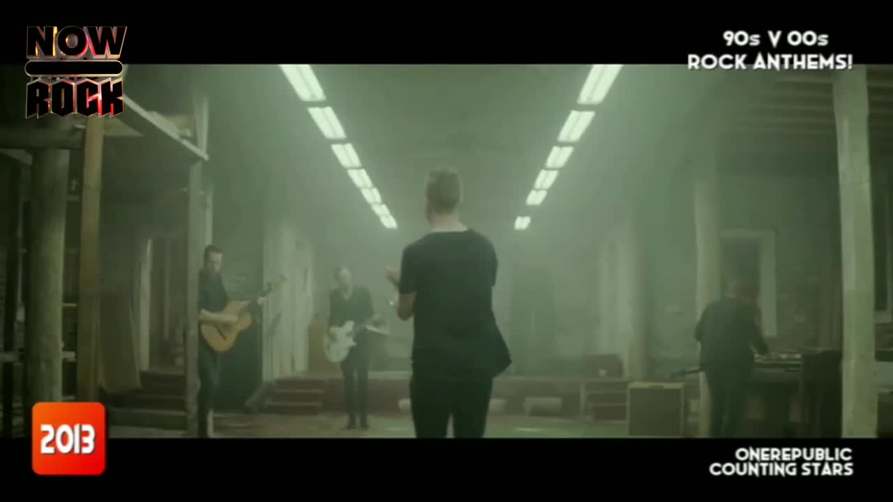 The lead singer, in a black shirt, stands center stage, facing away from the camera. Guitars and keyboards are positioned to either side, bathed in a hazy, green-tinged light.
