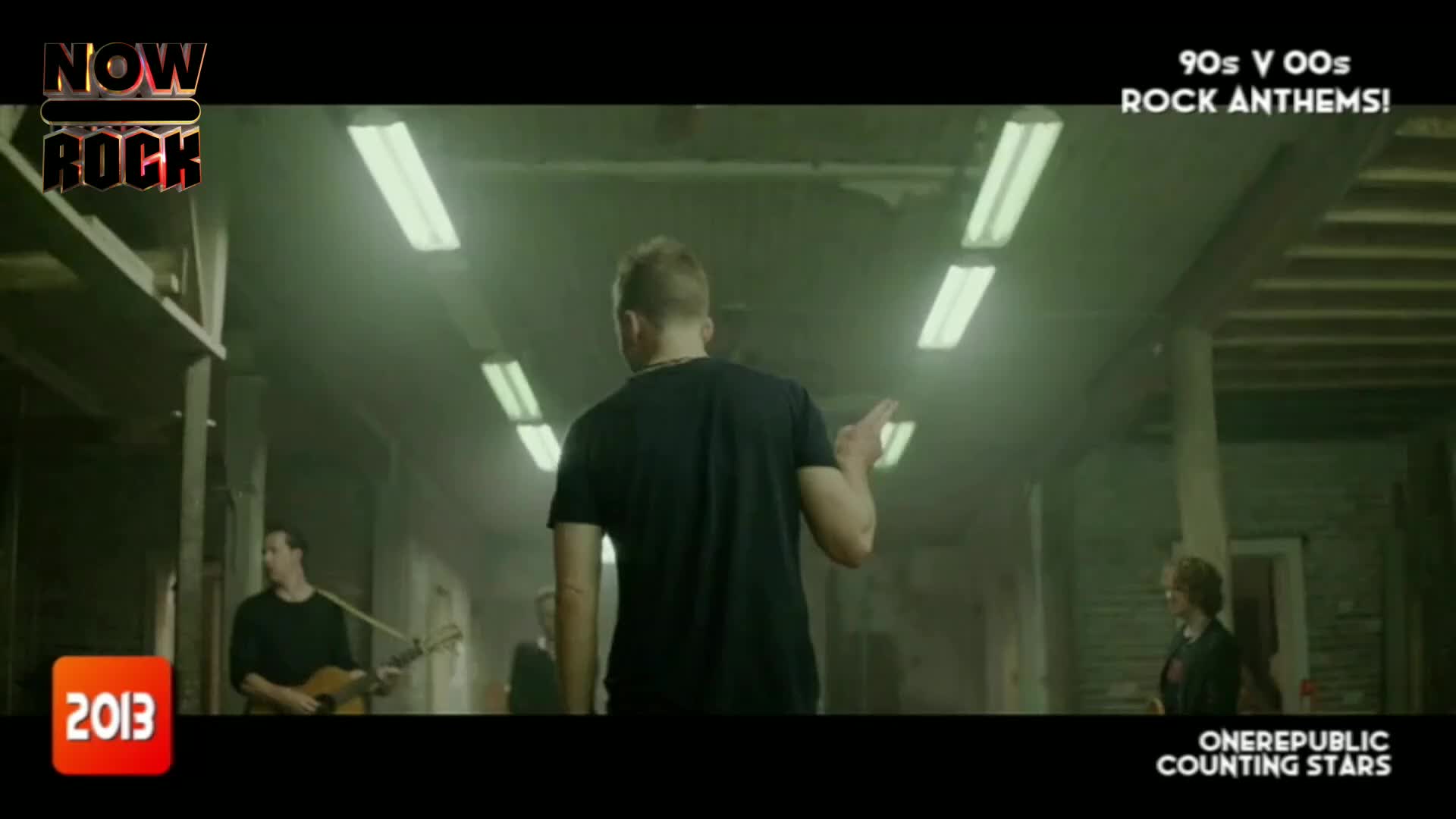A man in a black t-shirt gestures as he walks down a long, dimly lit corridor. Behind him, a band plays, illuminated by the overhead fluorescent lights.
A man in a black t-shirt gestures as he walks down a long, dimly lit corridor. Behind him, a band plays, illuminated by the overhead fluorescent lights.