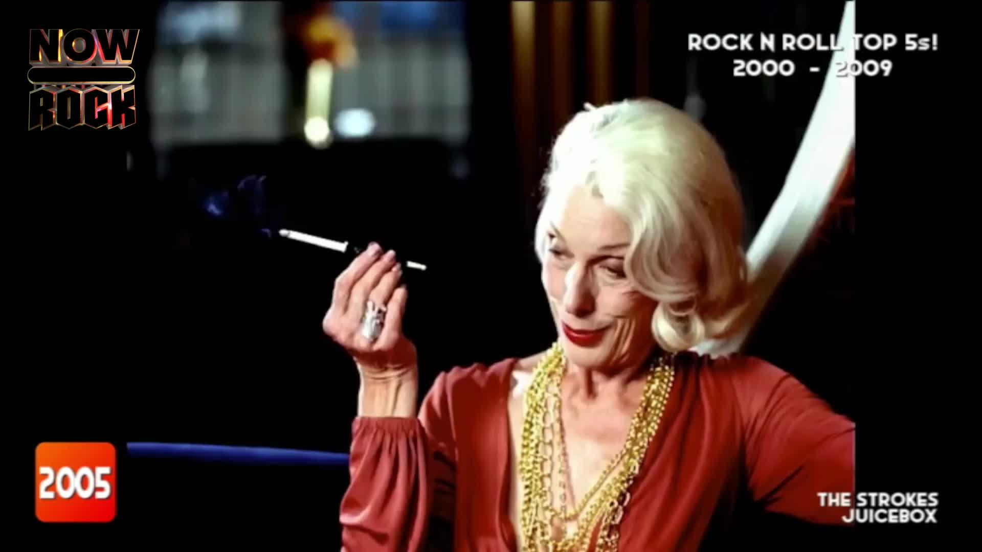 A woman with platinum hair and red lipstick holds a cigarette in a long holder, smoke curling upwards. She's wearing a red dress and a lot of gold necklaces, a vintage look for the NOW Rock countdown.
A woman with platinum hair and red lipstick holds a cigarette in a long holder, smoke curling upwards. She's wearing a red dress and a lot of gold necklaces, a vintage look for the NOW Rock countdown.