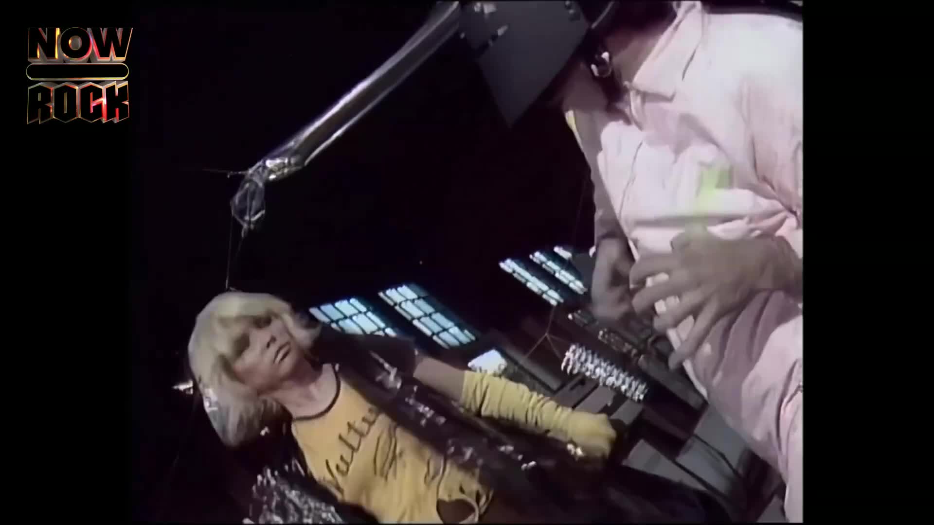 A woman in a yellow shirt and long gloves is lying down, playing a keyboard. A man in a pink suit is standing over her, working with a different keyboard.
A woman in a yellow shirt and long gloves is lying down, playing a keyboard. A man in a pink suit is standing over her, working with a different keyboard.