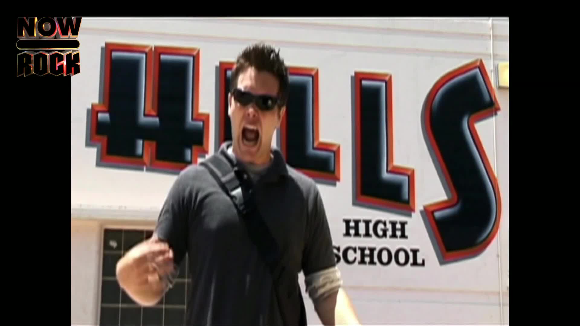A man in sunglasses stands in front of a building with "HILLS HIGH SCHOOL" written on it, shouting with his mouth wide open. He's gesturing with one hand as if he's making a point.
