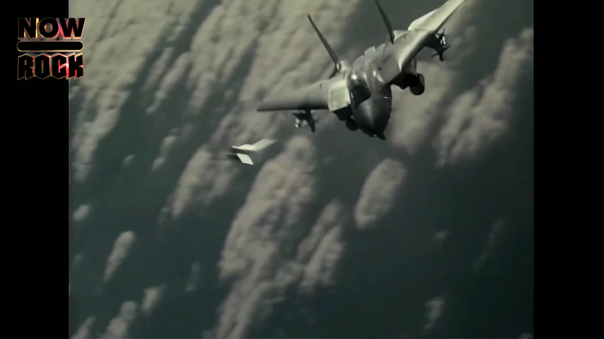 Two fighter jets, dark against the cloudy sky, bank sharply. One is closer, its angular design clearly visible, while the other trails behind.
