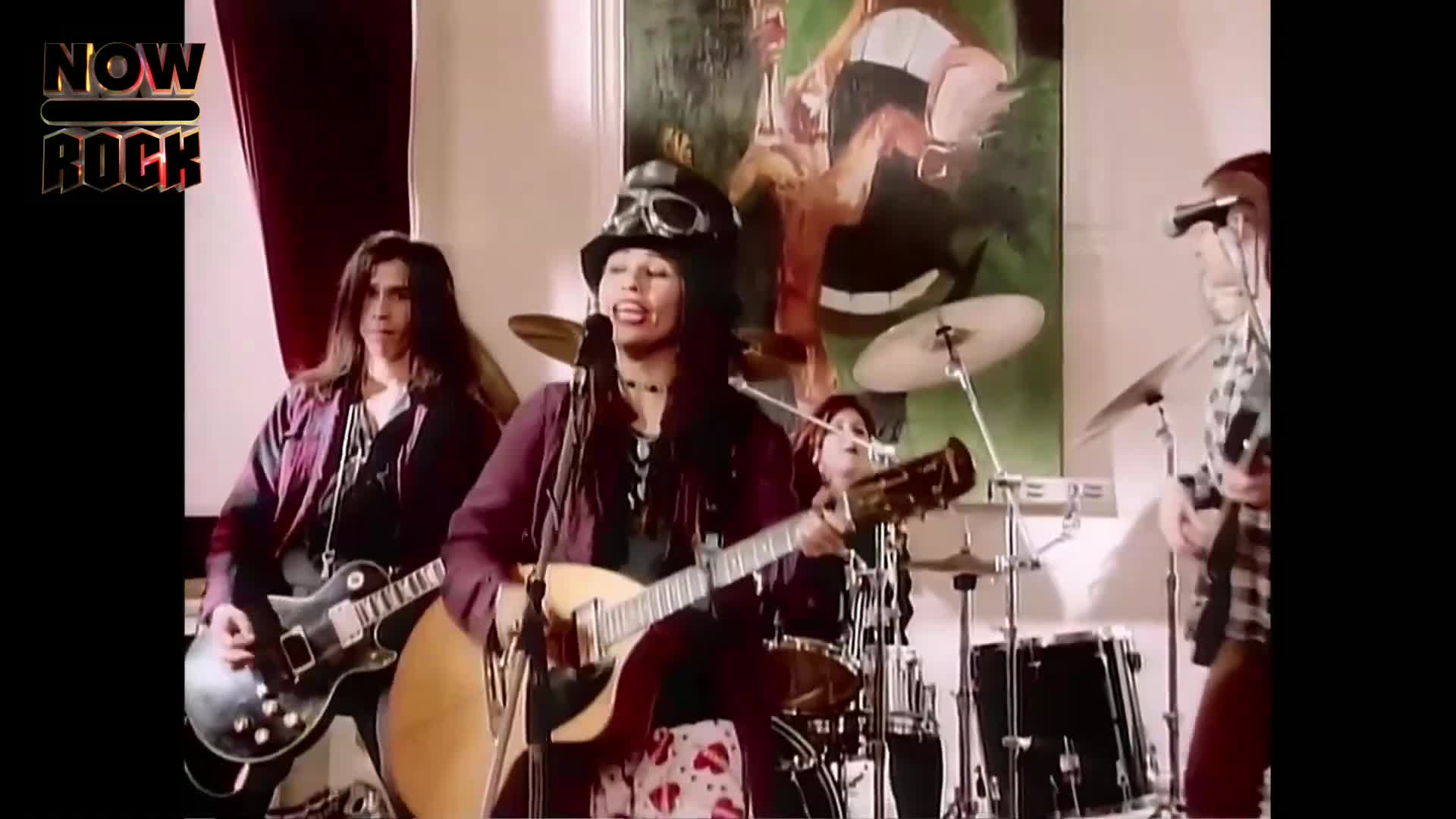 The lead singer, wearing a hat and goggles, strums an acoustic guitar and sings into a microphone. A band plays behind her, including a guitarist with long hair, a drummer, and another guitarist.
