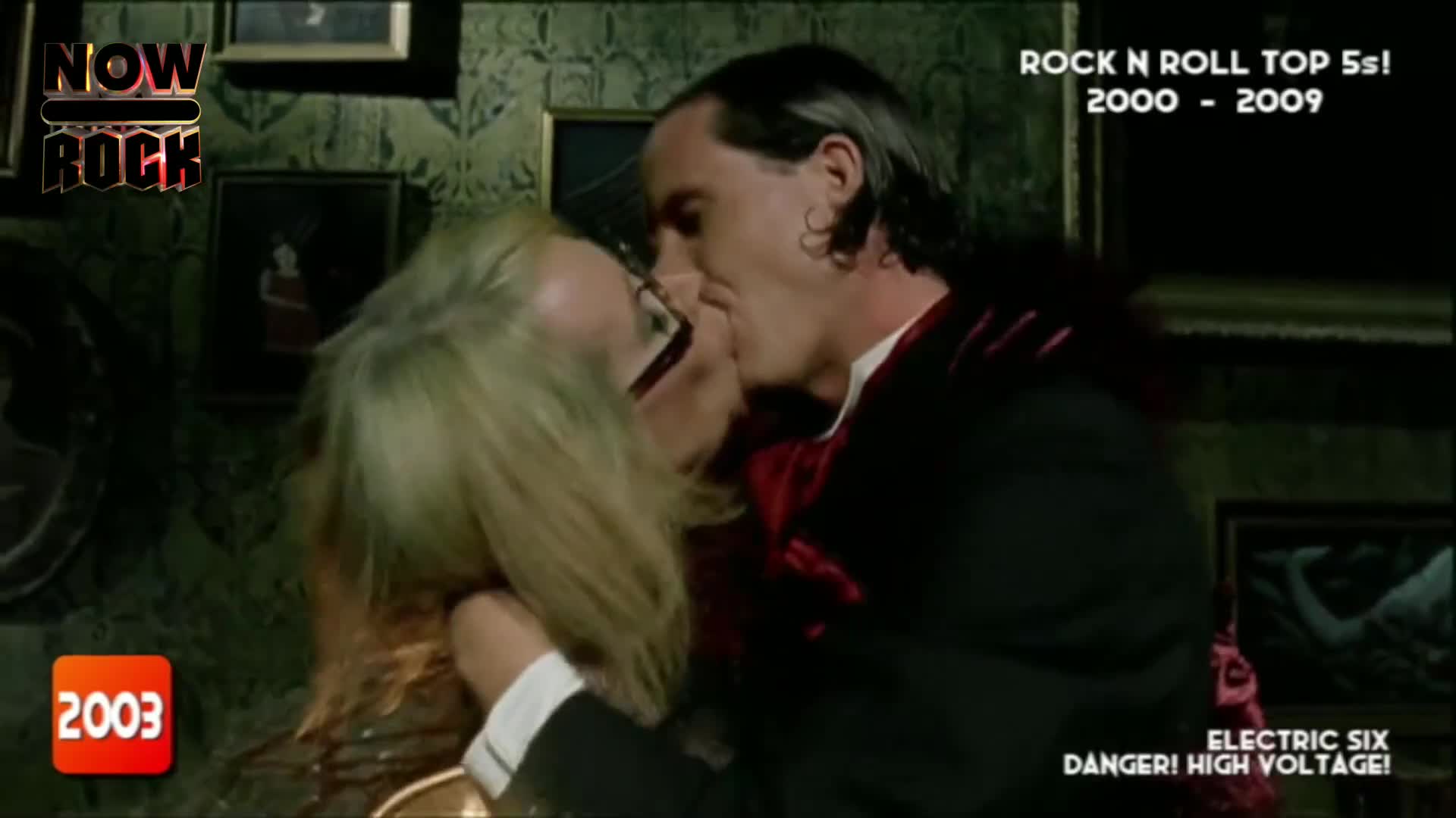A man in a black suit and a woman with blonde hair are locked in a kiss. The scene looks like a music video from the early 2000s, with a "NOW Rock" logo in the corner.
