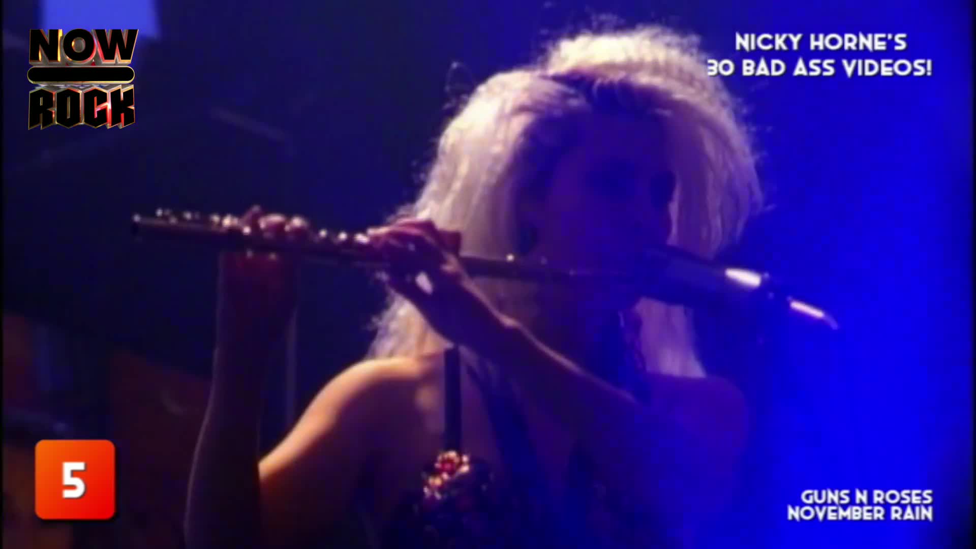 A woman with bright blonde hair holds a flute, her face partially obscured by the instrument and the stage lighting. The NOW Rock logo and a countdown number are visible, suggesting a music video from the UK.

