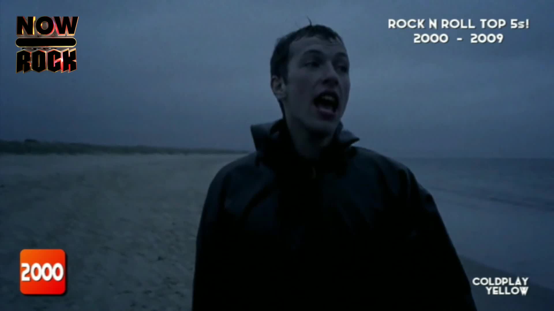 A man, drenched in a dark jacket, is running towards the camera on a windswept beach. His mouth is wide open, mid-scream, as he faces the grey sky and the churning sea.
