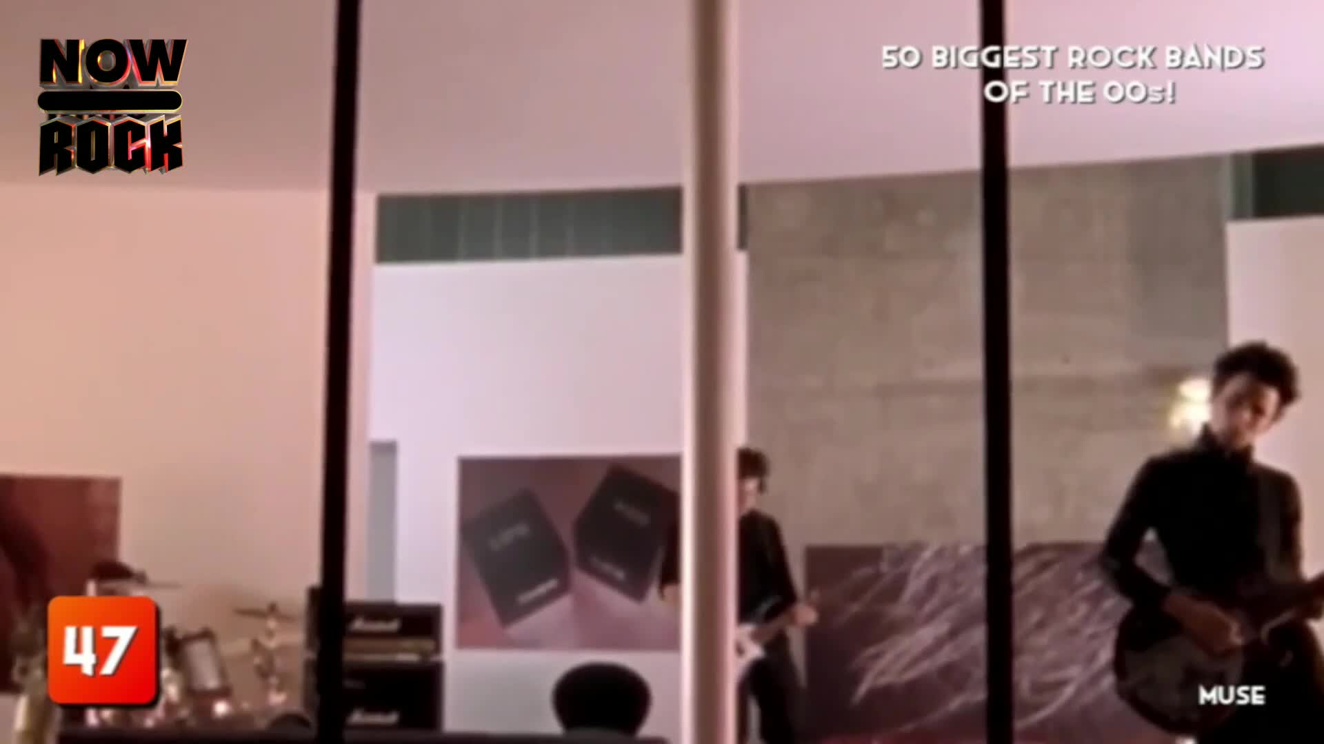 A guitarist in a dark suit strums a black guitar, the light catching his face. The scene is part of a NOW Rock countdown, with the band Muse featured in the background.
