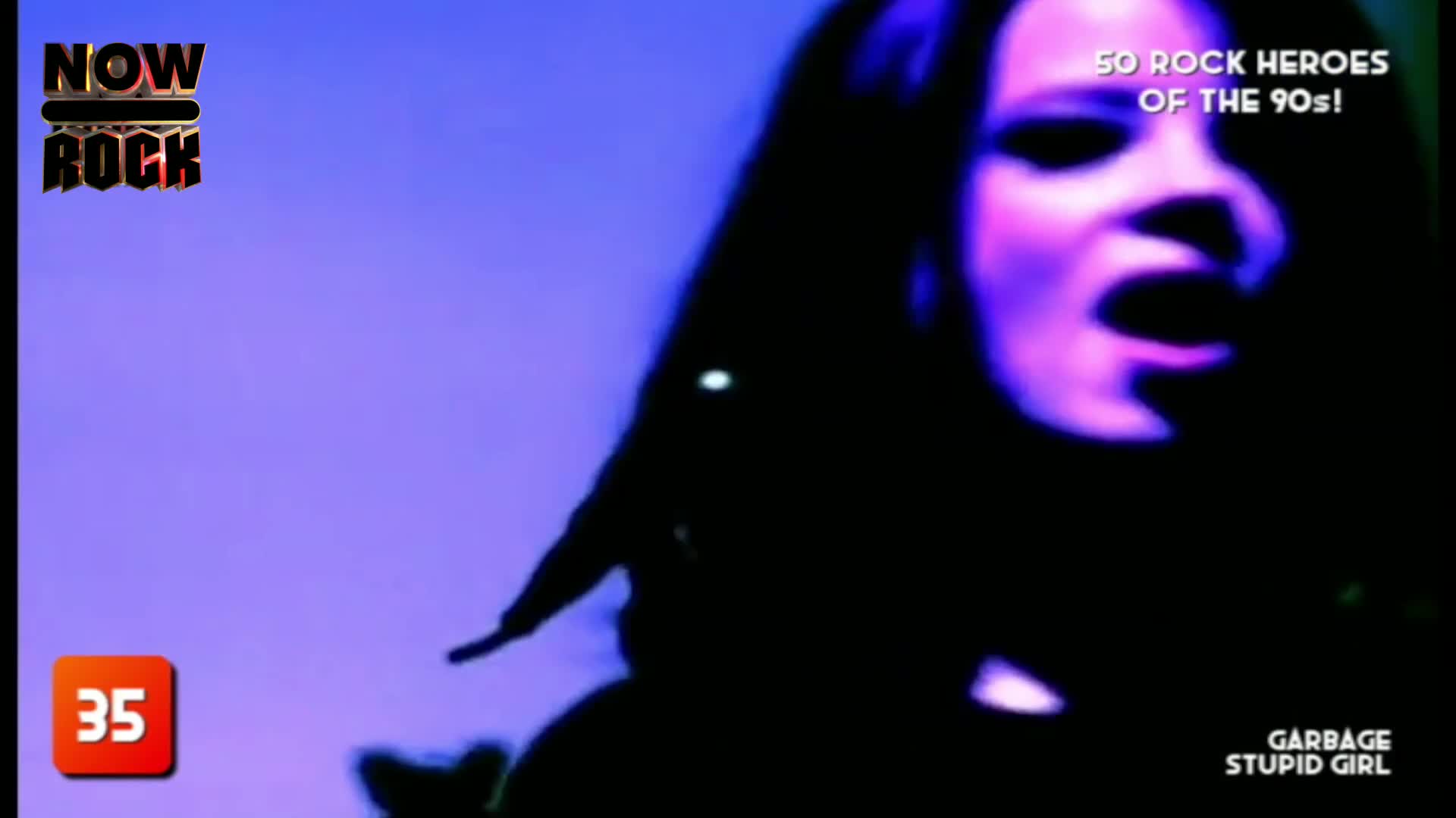 The singer's face is lit with purple, her mouth wide open as she belts out a song. Long, dark hair frames her face, and the background is a solid, cool-toned blue.
