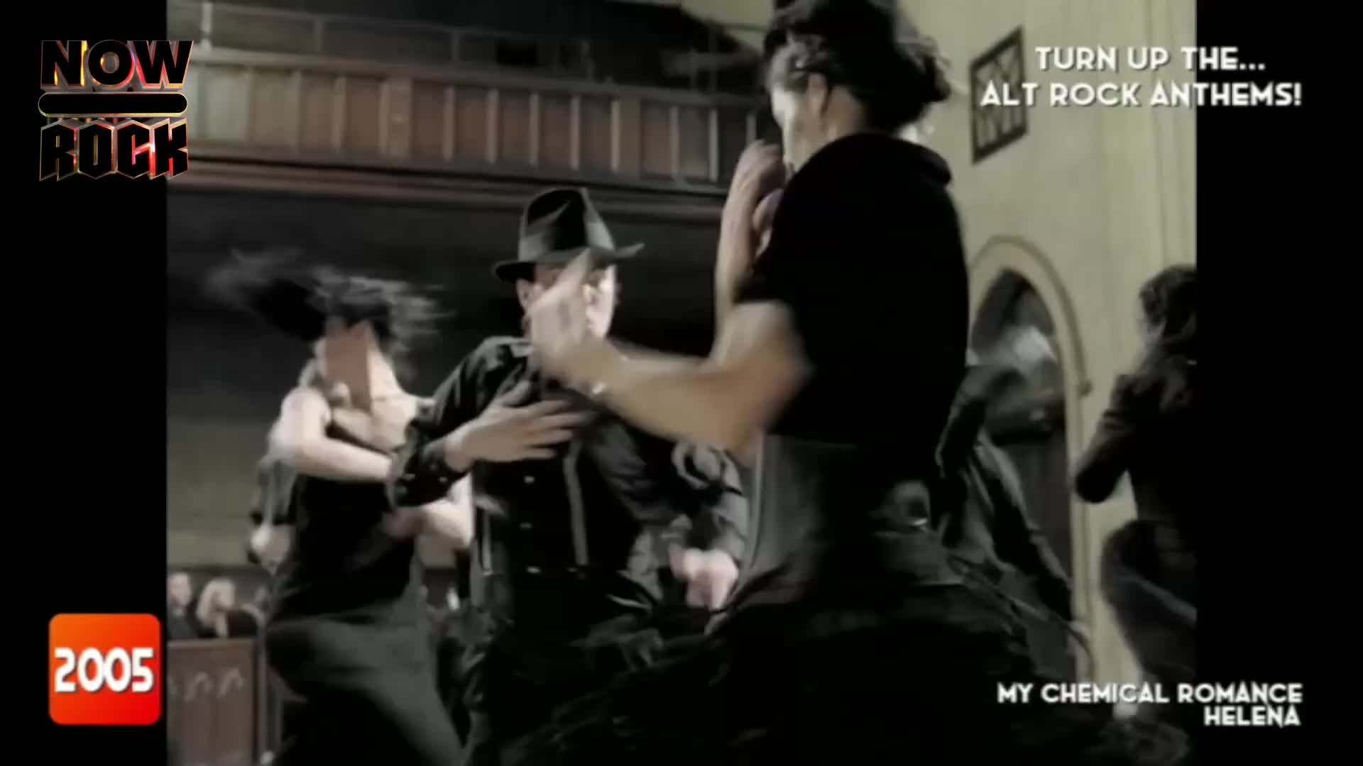 Dancers in dark clothing move with jerky, stylized motions in a dimly lit space. The lead singer, wearing a fedora, gestures with his hands as the song builds.
