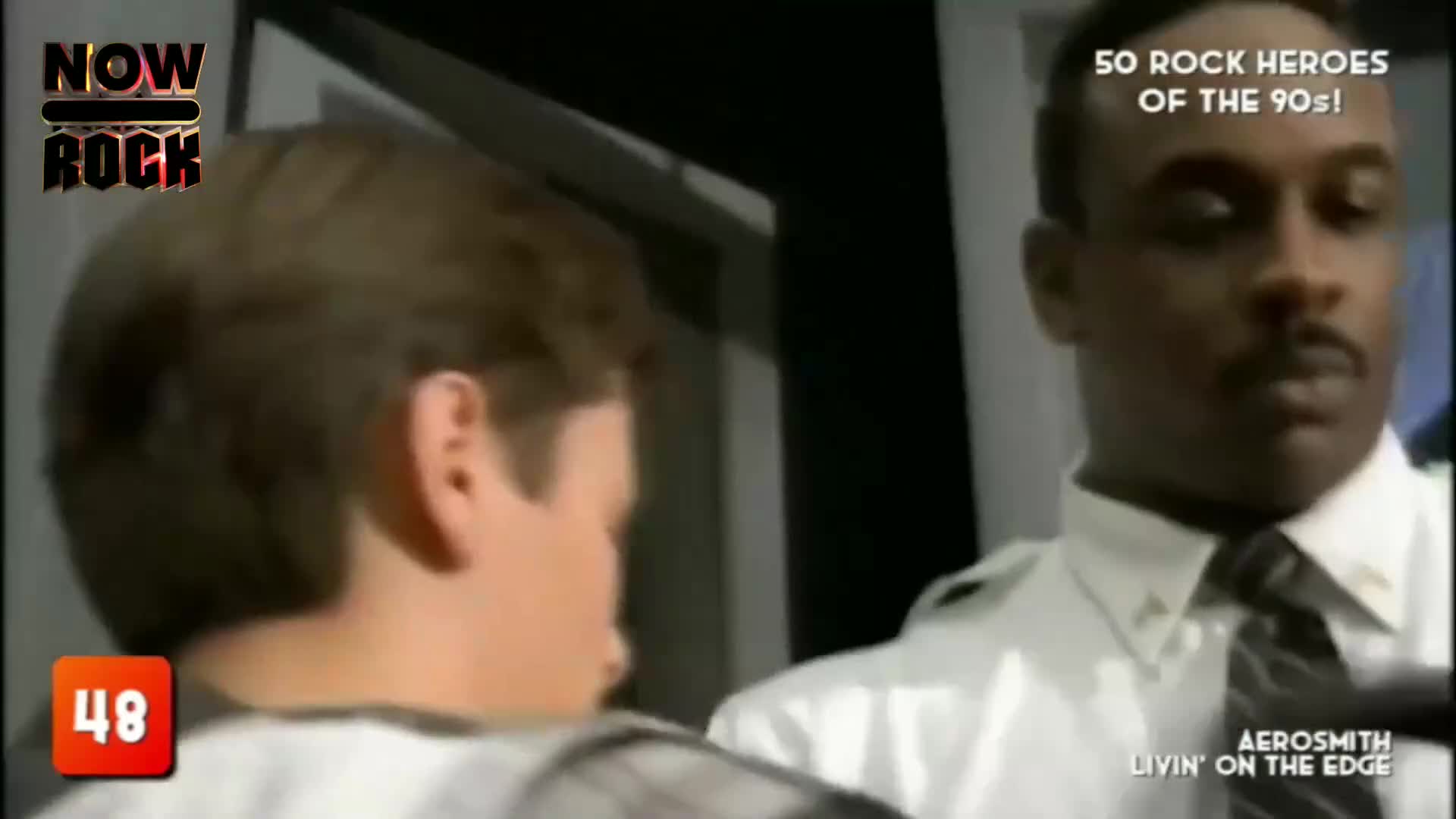 A man in a white shirt and tie stands close to a younger man, both facing the same direction. The "NOW Rock" logo and the "50 Rock Heroes of the 90s!" text suggest this is a clip from a music program.
