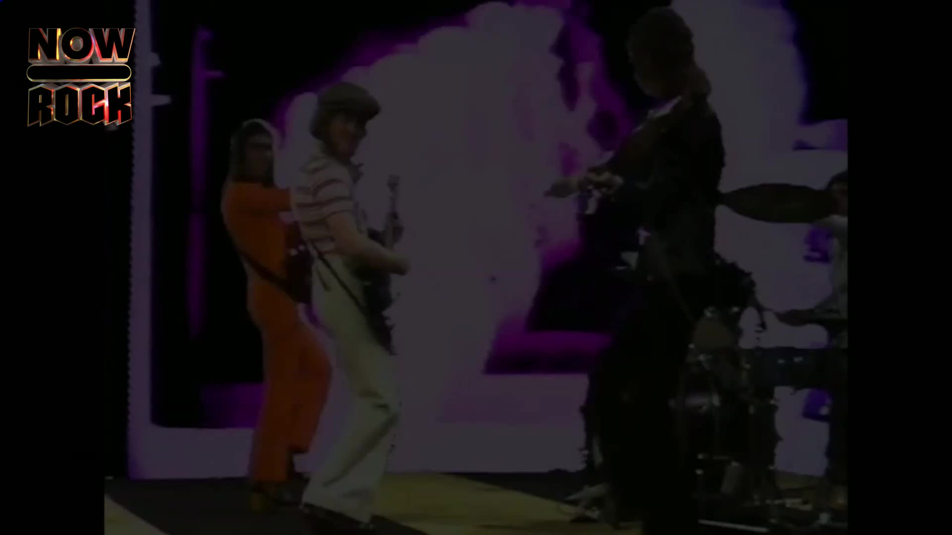 A guitarist in a striped shirt and cap plays his instrument, while another band member in an orange jumpsuit stands beside him. The stage lights cast a purple glow behind them. A guitarist in a striped shirt and cap plays his instrument, while another band member in an orange jumpsuit stands beside him. The stage lights cast a purple glow behind them.