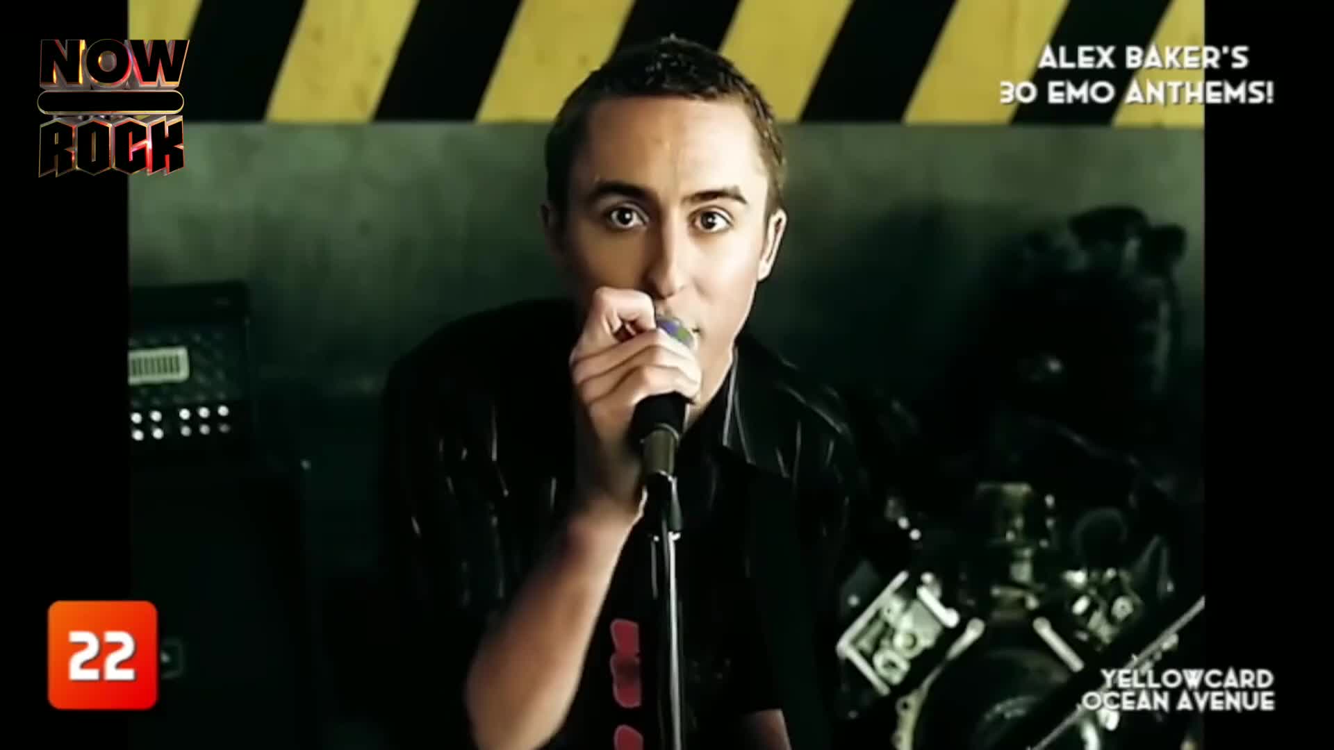 A young man with wide eyes grips a microphone, singing into it. Behind him, a dark industrial space is lit by yellow and black hazard stripes on the wall.