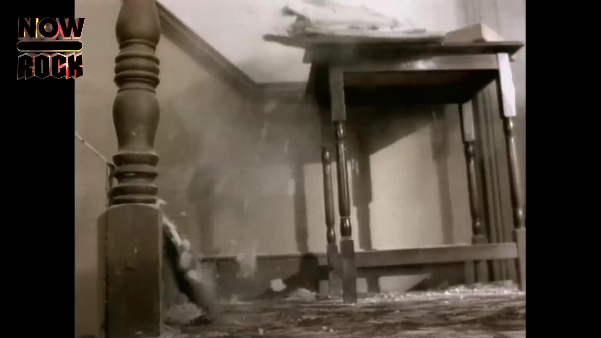 Dust and debris are falling from the wall. A wooden table stands to the right, its legs casting shadows.