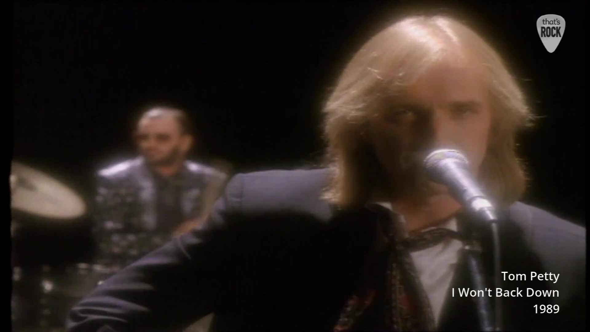 The singer leans into the microphone, his blonde hair catching the light. Behind him, a drummer is visible, his hands a blur over the cymbals. The singer leans into the microphone, his blonde hair catching the light. Behind him, a drummer is visible, his hands a blur over the cymbals.
