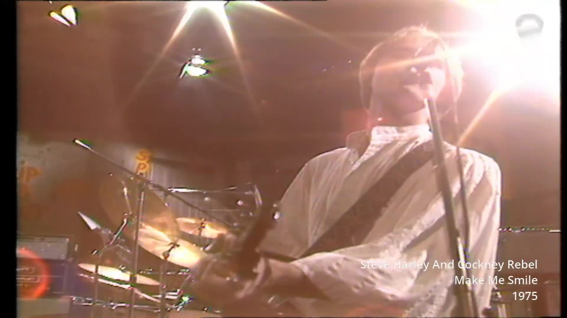 The lead singer of Cockney Rebel, Steve Harley, belts into the microphone, his white shirt catching the stage lights. Behind him, the drummer hits a cymbal, the metal glinting. The lead singer of Cockney Rebel, Steve Harley, belts into the microphone, his white shirt catching the stage lights. Behind him, the drummer hits a cymbal, the metal glinting.