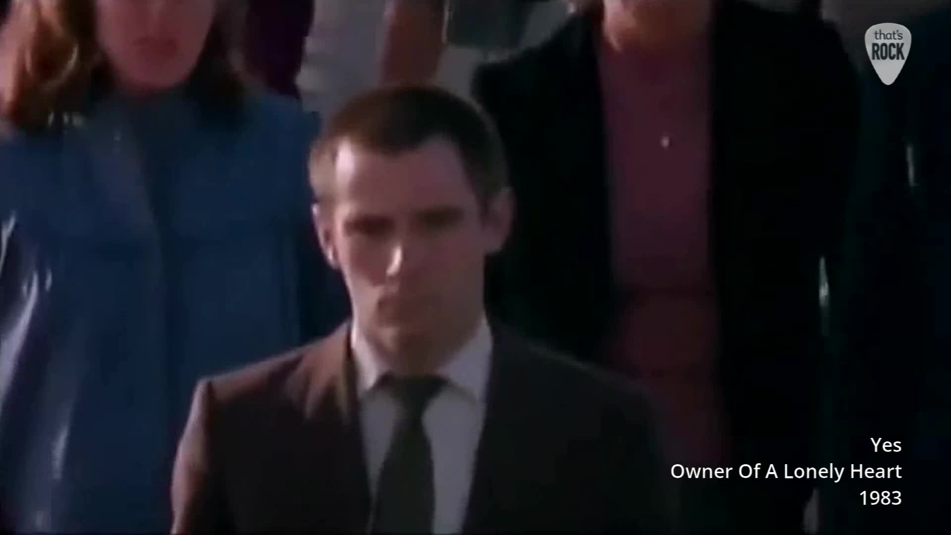 A man in a suit walks forward, his expression serious. He's surrounded by others in what looks like a crowd at a concert. A man in a suit walks forward, his expression serious. He's surrounded by others in what looks like a crowd at a concert.