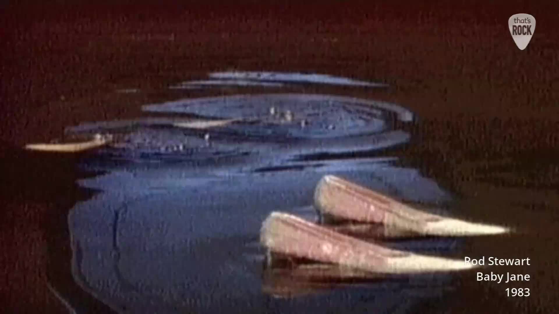 Two pairs of pointed, pale pink shoes float just beneath the surface of dark, rippling water. A faint yellow object drifts on the left, leaving a subtle wake. Two pairs of pointed, pale pink shoes float just beneath the surface of dark, rippling water. A faint yellow object drifts on the left, leaving a subtle wake.