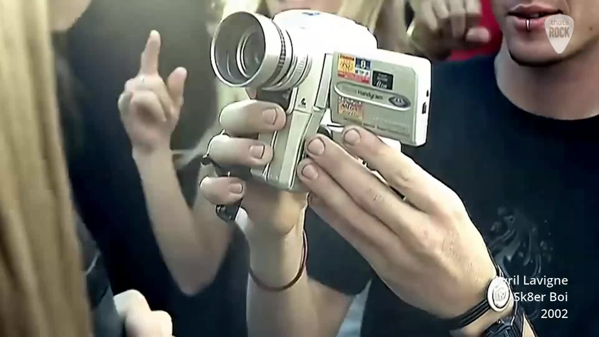 A hand steadies a camcorder, its lens pointed forward. Behind it, another person gestures with a raised finger. A hand steadies a camcorder, its lens pointed forward. Behind it, another person gestures with a raised finger.