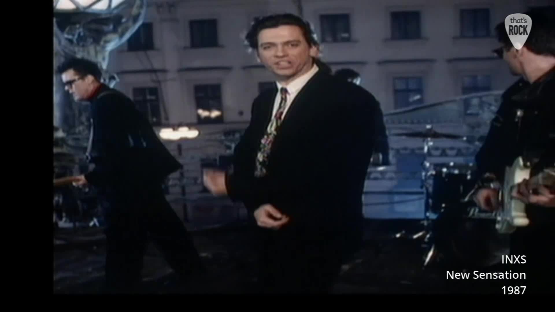 The band is playing on a city street at night, the lead singer in a sharp suit and colourful tie is animatedly singing. A guitarist on the left, also in a suit, plays his instrument as the drummer can be glimpsed behind them. The band is playing on a city street at night, the lead singer in a sharp suit and colourful tie is animatedly singing. A guitarist on the left, also in a suit, plays his instrument as the drummer can be glimpsed behind them.