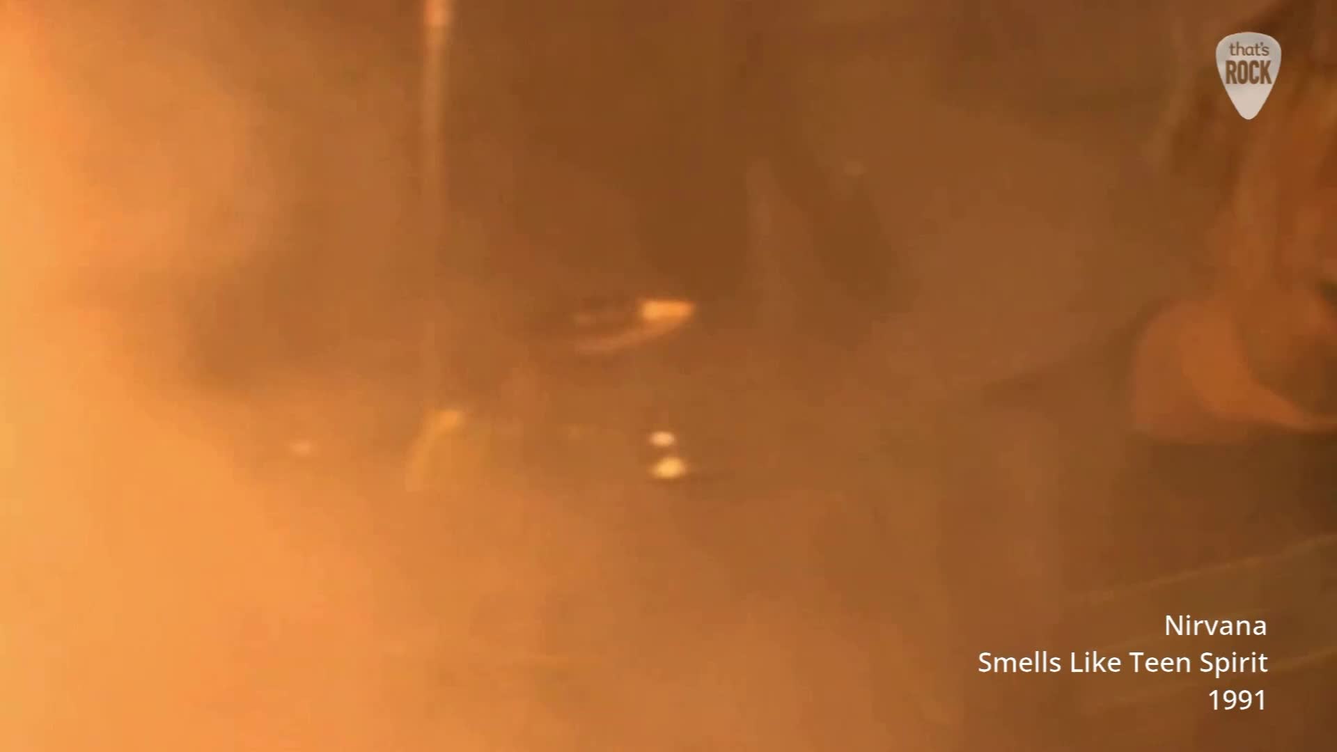 Smoke billows around the band as they play, the stage lights casting an orange glow. A guitar neck is visible, held aloft by a blurred figure. Smoke billows around the band as they play, the stage lights casting an orange glow. A guitar neck is visible, held aloft by a blurred figure.