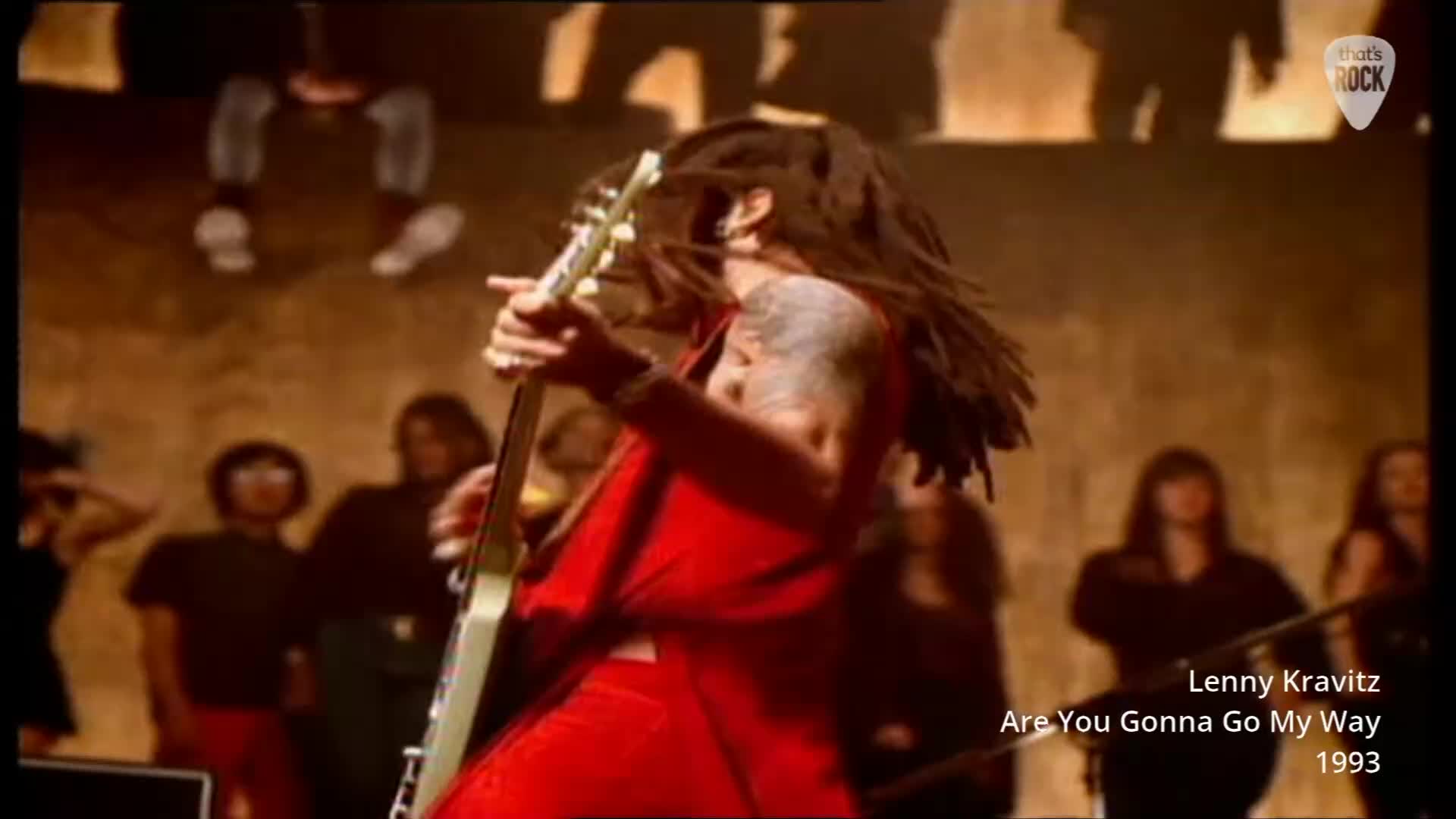Lenny Kravitz is thrashing his guitar, his dreadlocks flying as he performs. The crowd behind him watches intently, bathed in warm stage lights.