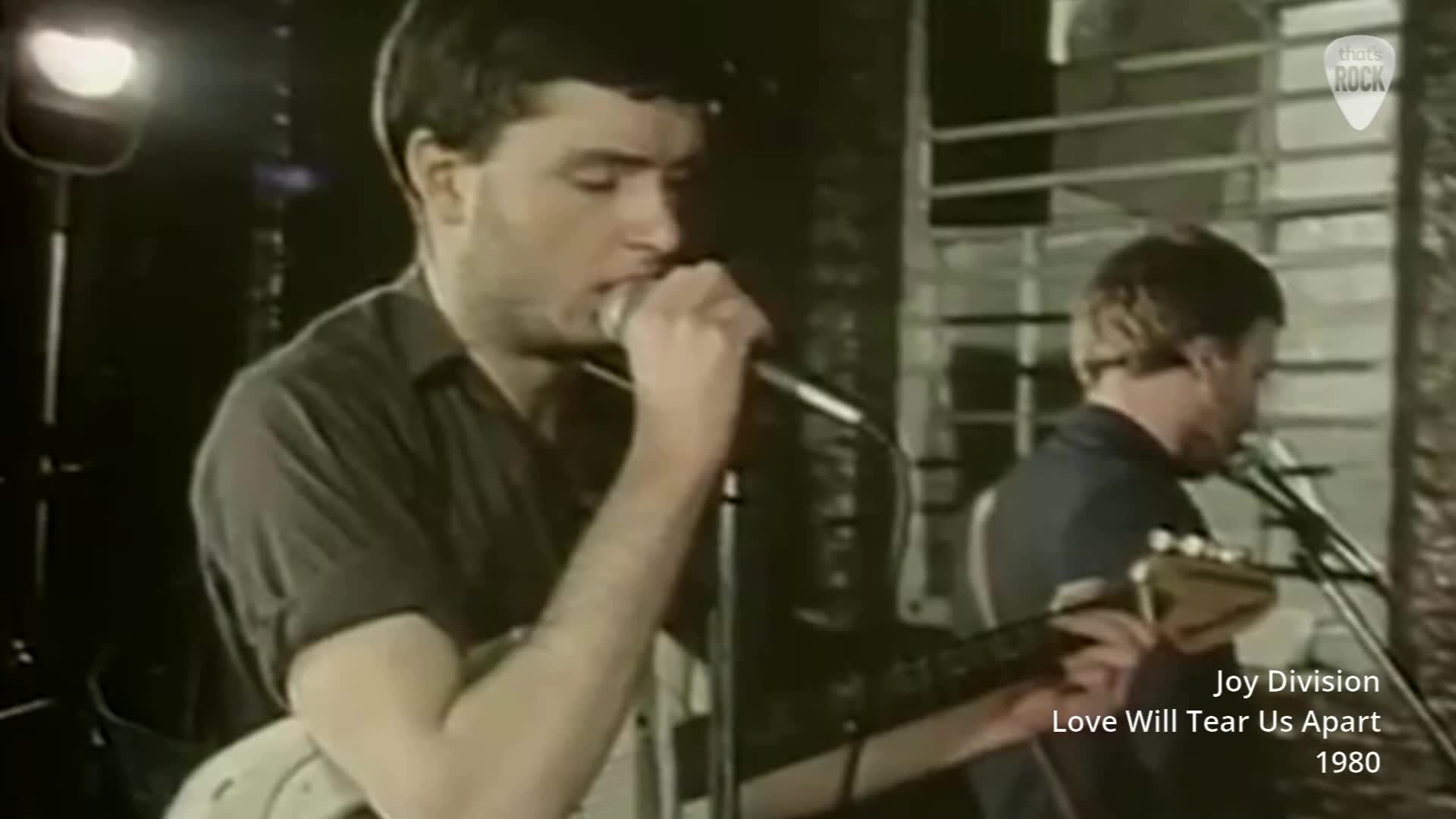 A man with dark hair sings into a microphone, his guitar resting on his shoulder. Another man plays guitar behind him, his face turned away.