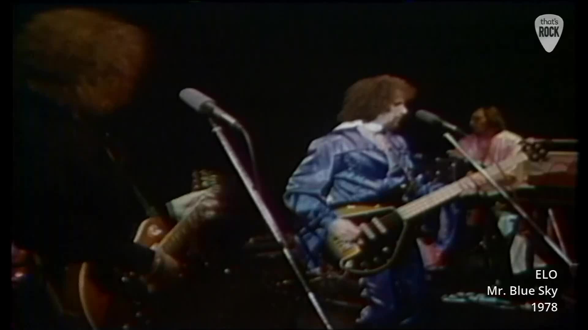 A bassist in a shiny blue jumpsuit plays his instrument, his head tilted as he sings into the microphone. To his left, a guitarist with a mop of brown hair leans into his own mic, his fingers a blur on the fretboard.