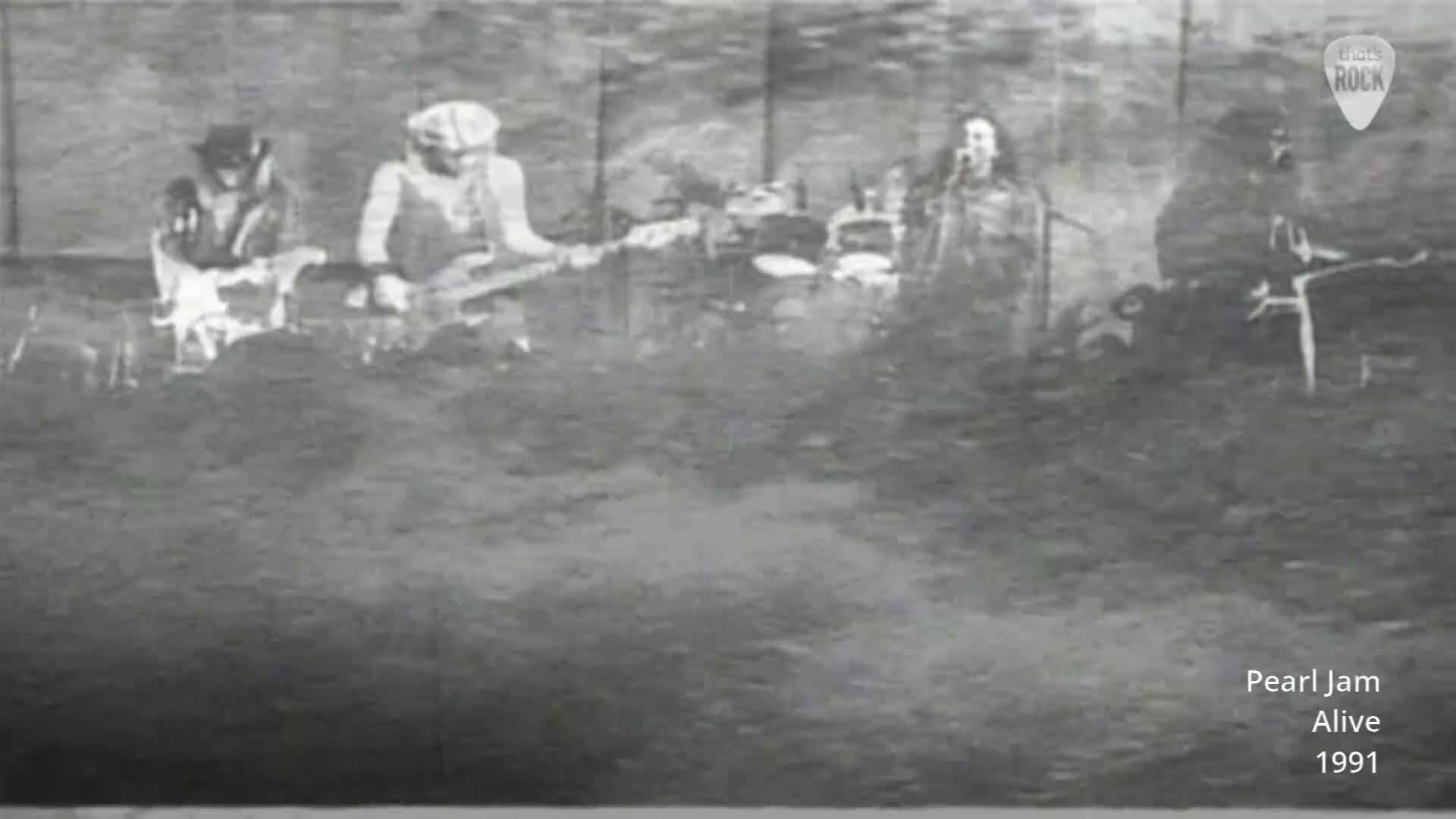 A band plays on a stage, bathed in what looks like a hazy spotlight. The guitarist on the left wears a hat, while the bassist in the center is clad in a beanie.