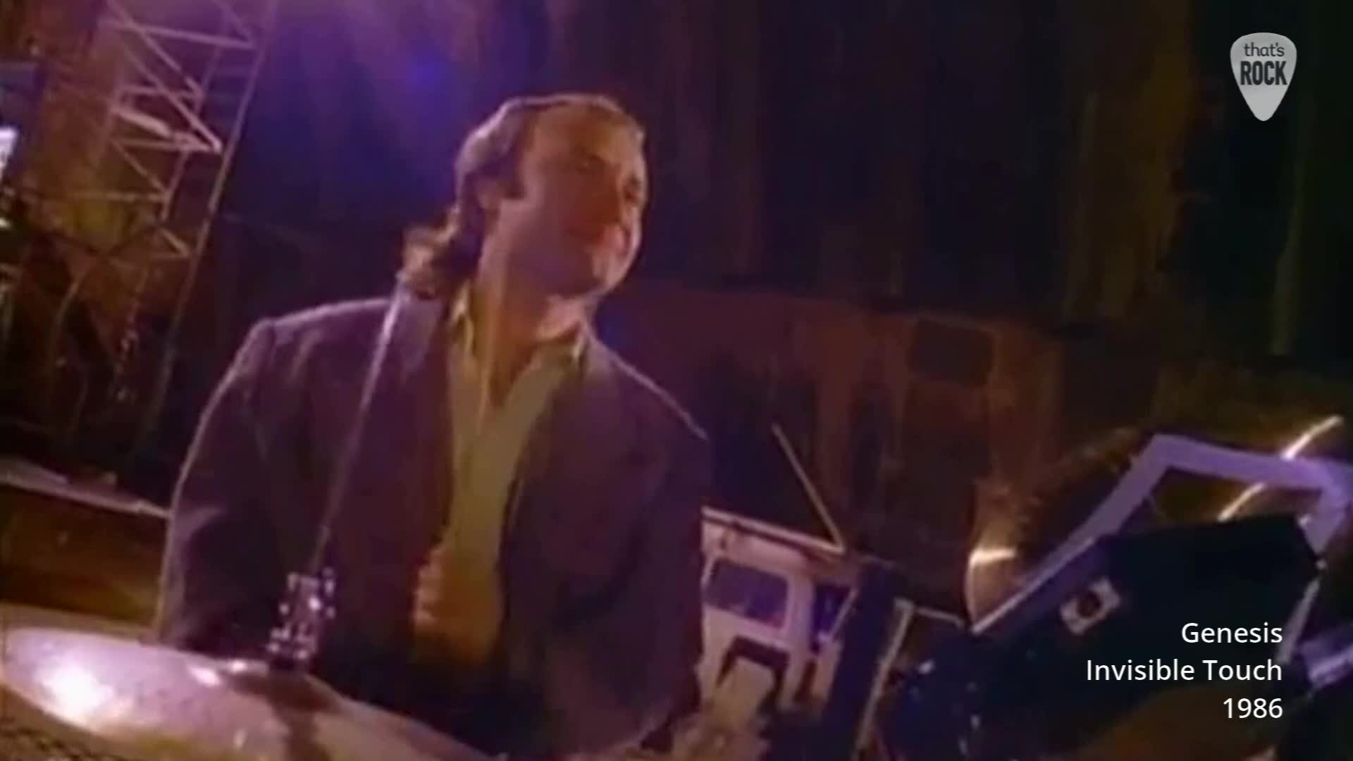 Phil Collins is playing the drums, his arms a blur as he hits the cymbals. The stage lights cast a warm glow on the band as they perform.