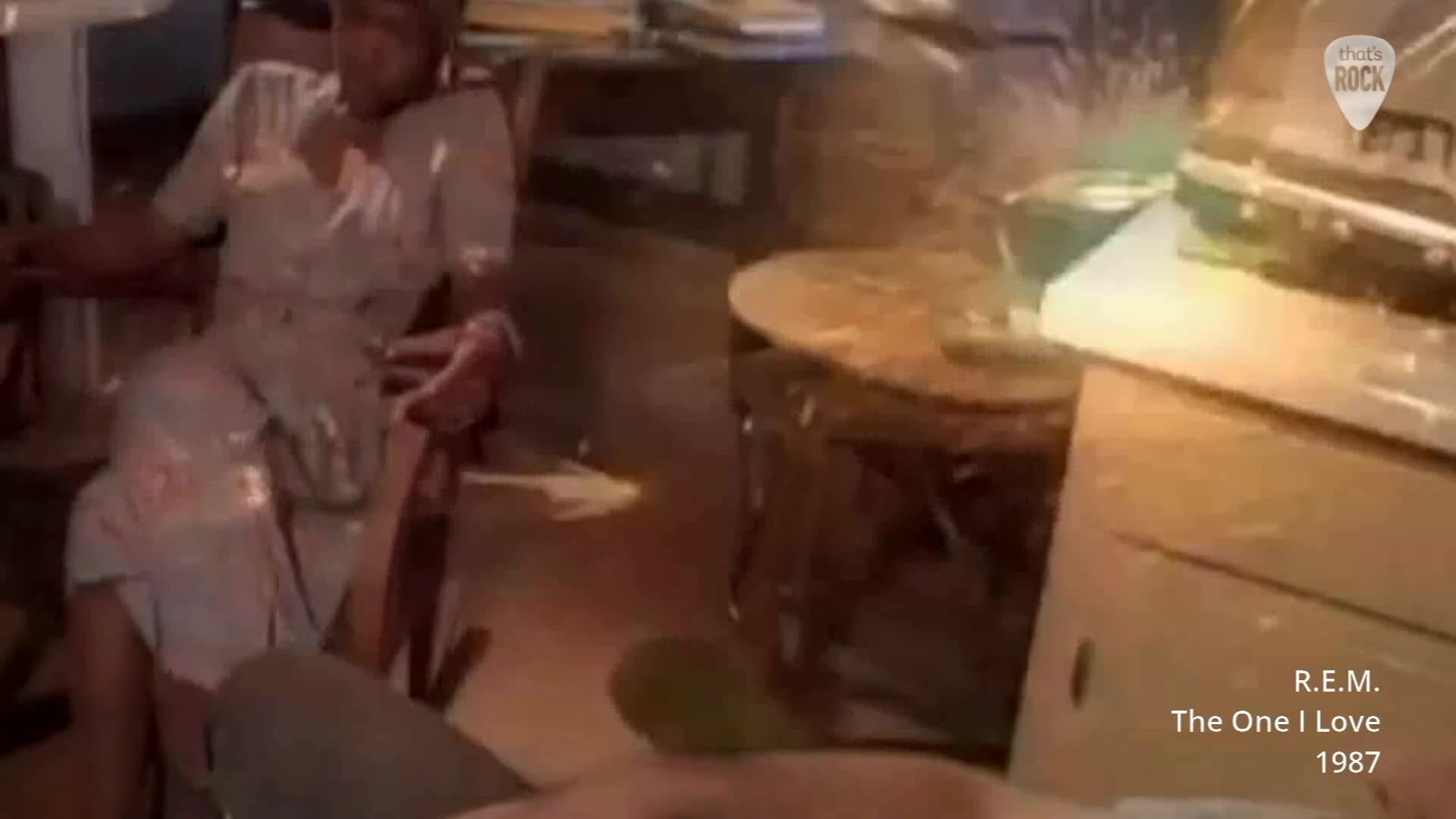 A woman in a light-coloured dress sits in a wooden chair, her head tilted back. The room is dimly lit, with a round table and what looks like a record player in the background.