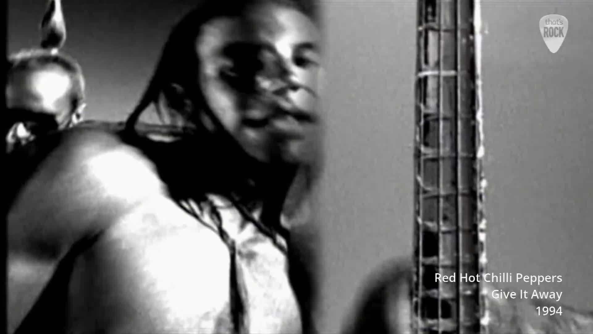 A shirtless man with long hair is playing a bass guitar, his face a blur of motion. Another man, his head shaved, stands behind him, his gaze fixed forward.