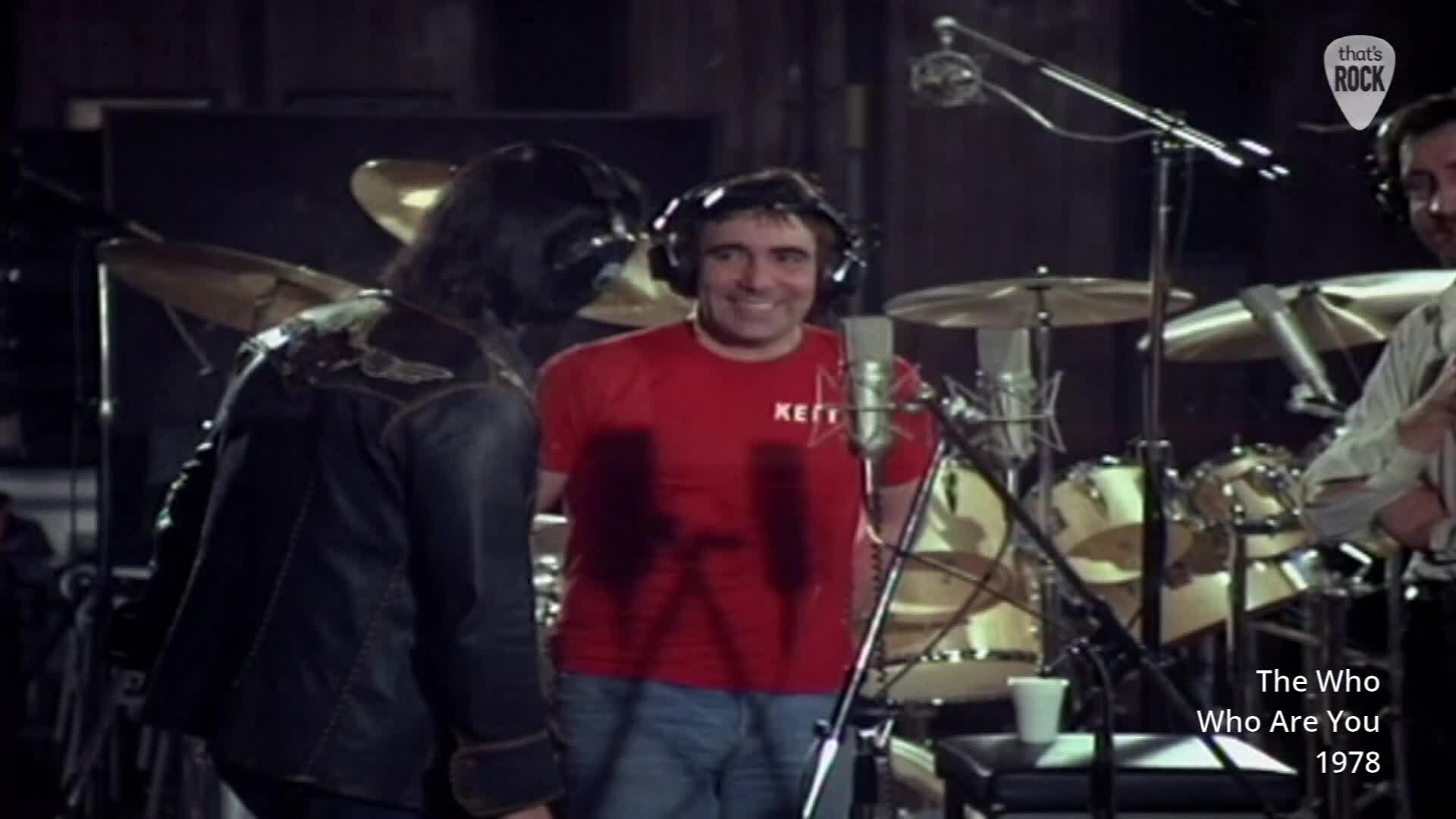 A man in a red t-shirt grins, wearing headphones. Beside him, another man in a dark leather jacket stands by a drum kit.