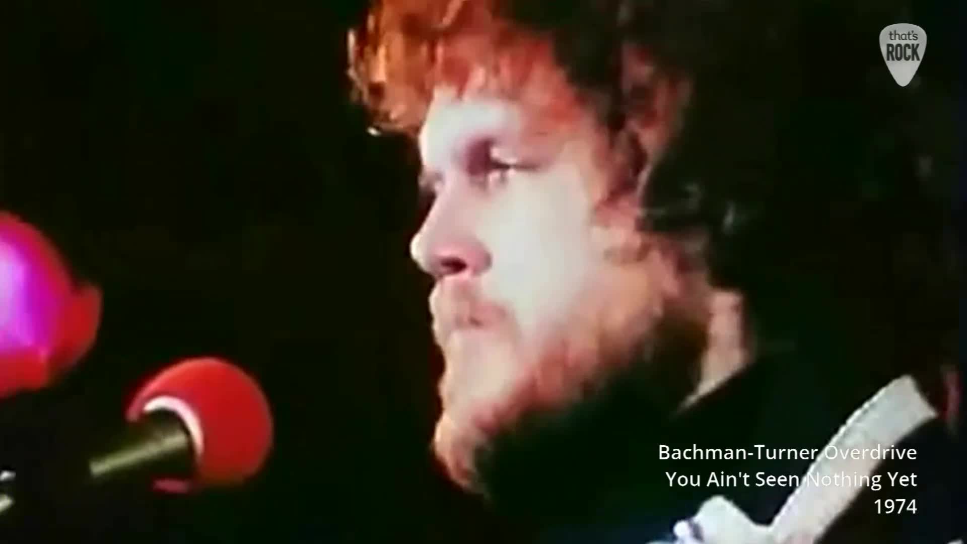 The singer's face, framed by auburn hair, is turned away from the audience, his gaze fixed on something unseen. Two microphones stand ready, their red foam heads catching the stage lights.