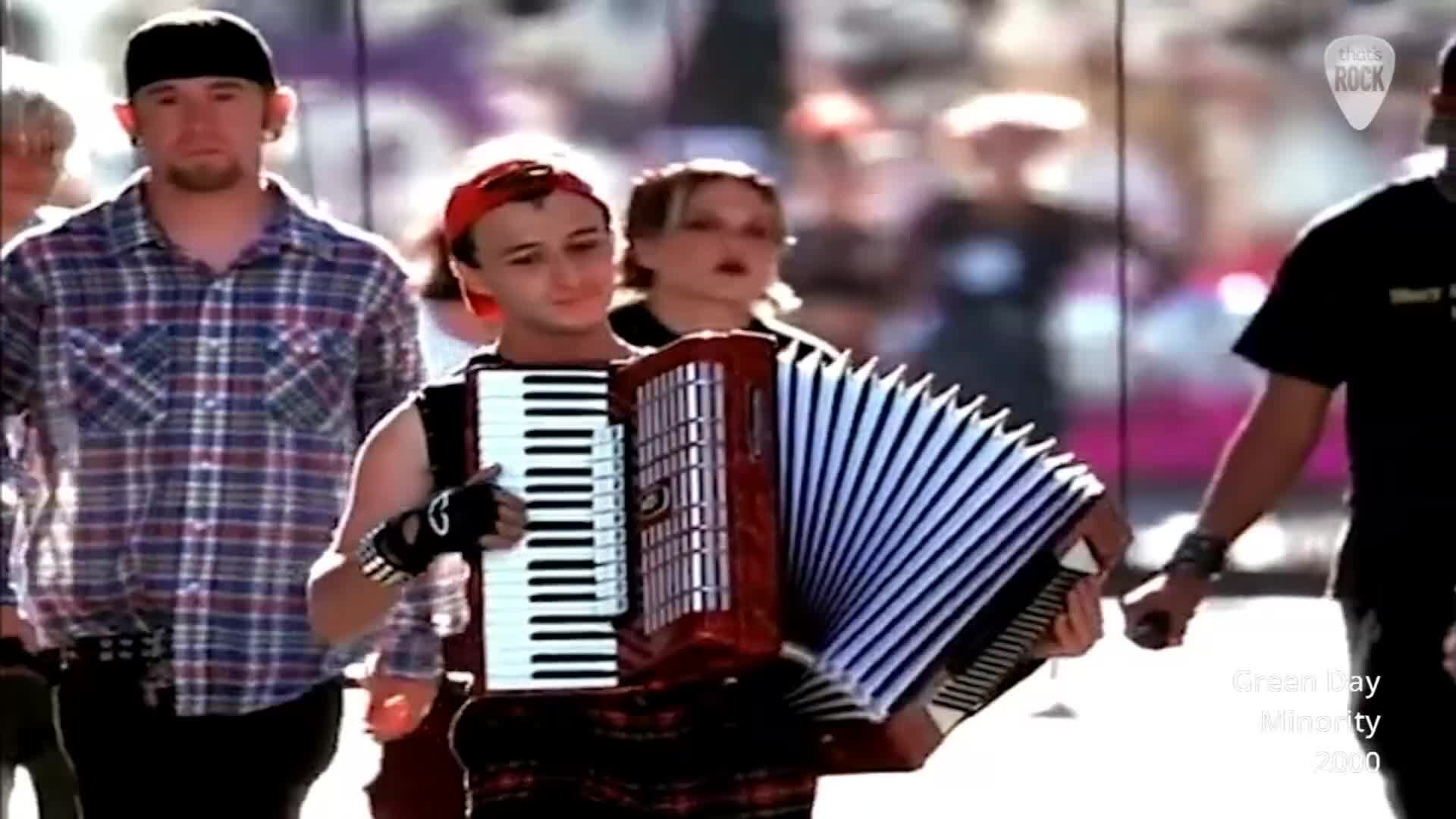 A young person in a red bandana plays an accordion, their fingers moving across the keys. Behind them, a man in a plaid shirt walks with a determined expression.