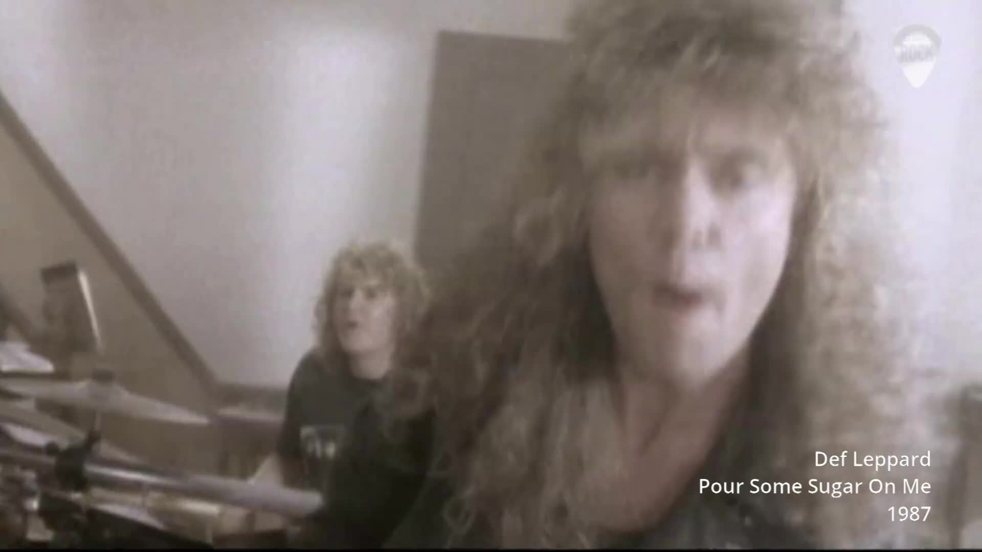 The singer leans into the camera, his mouth open mid-song. Behind him, the drummer is a blur of motion at his kit.