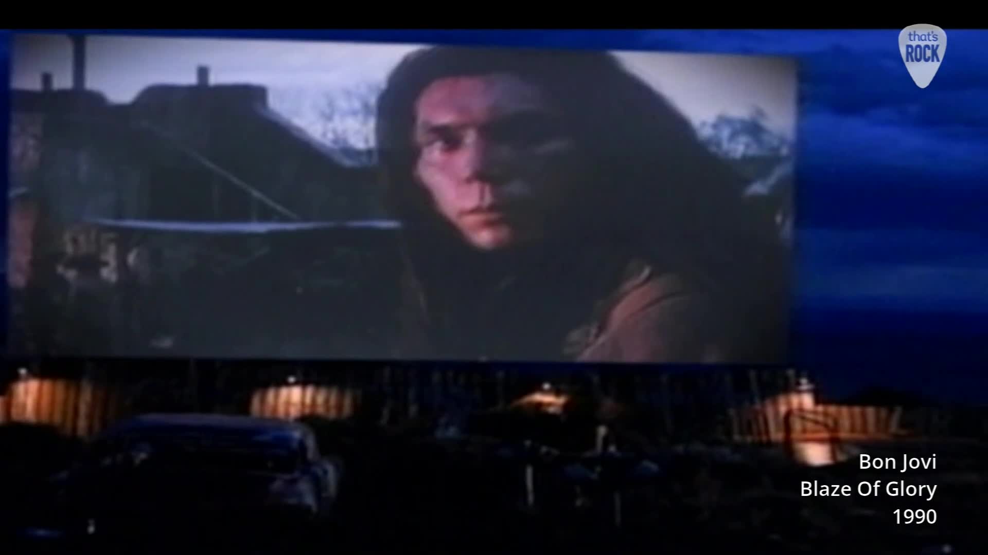 A young man with long hair stares intently from a large screen, the grainy footage suggesting a classic rock video. Below, a few cars are parked at a drive-in theatre, their silhouettes dark against the illuminated screen.