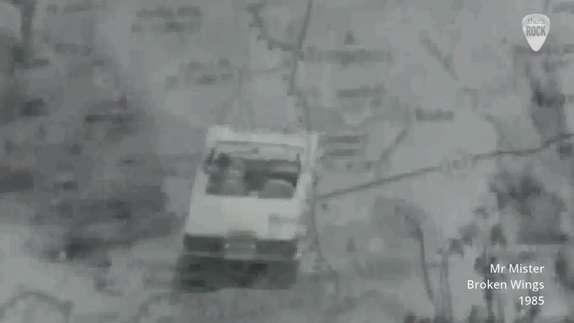 A white convertible drives along a road that cuts through a grey landscape. The car, with its top down, appears to be heading towards a junction.