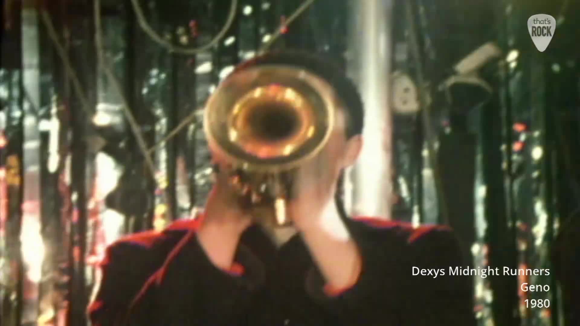 A man in a dark jacket plays a brass horn, his face obscured by the instrument. Behind him, shimmering silver curtains catch the stage lights.