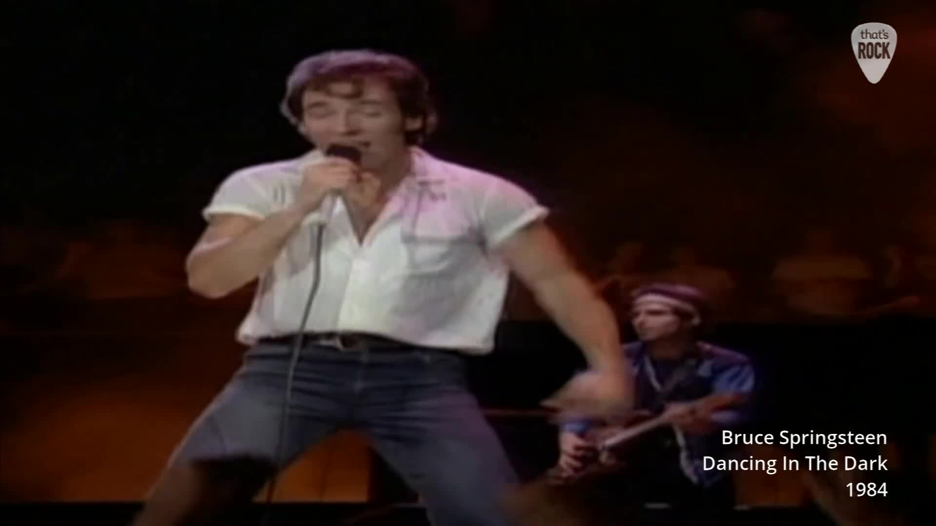 Bruce Springsteen belts into the microphone, his white shirt sleeves rolled up. Behind him, a guitarist is a blur of motion, lost in the music.