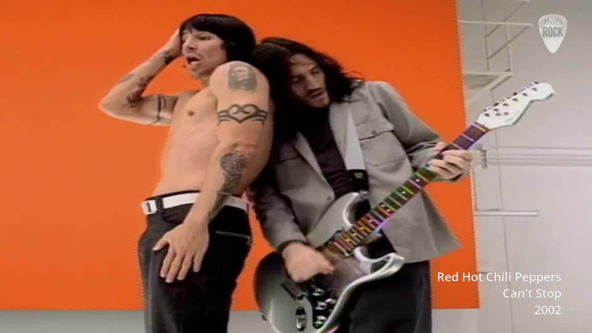 The guitarist leans into the singer, his hand strumming the rainbow-fretted guitar. The singer, bare-chested, raises a hand to his forehead, his gaze directed upwards.