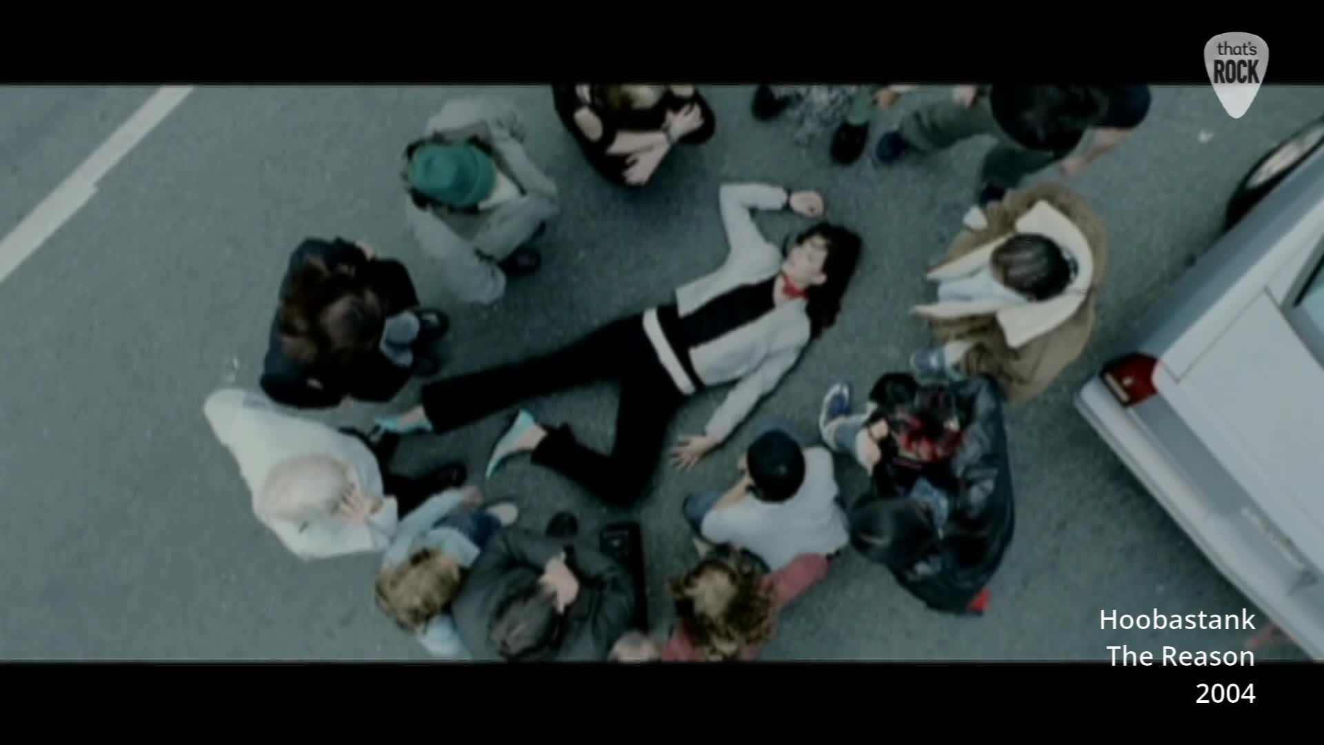 A young man lies sprawled on the tarmac, surrounded by a circle of onlookers. A silver car is parked nearby, its door open. A young man lies sprawled on the tarmac, surrounded by a circle of onlookers. A silver car is parked nearby, its door open.