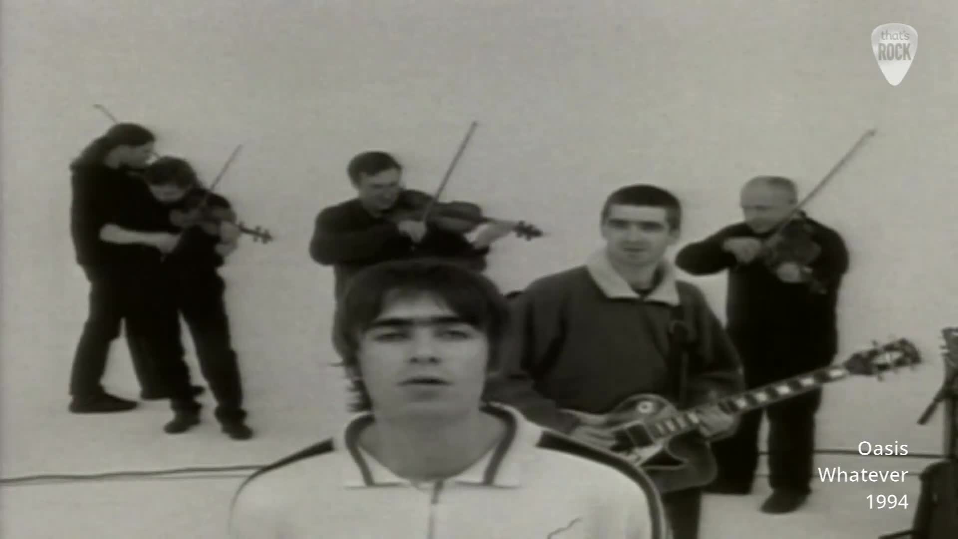 A man with a guitar stands in front of a white backdrop, while four violinists play behind him. The lead singer, Liam Gallagher, is at the forefront, looking directly ahead. A man with a guitar stands in front of a white backdrop, while four violinists play behind him. The lead singer, Liam Gallagher, is at the forefront, looking directly ahead.
